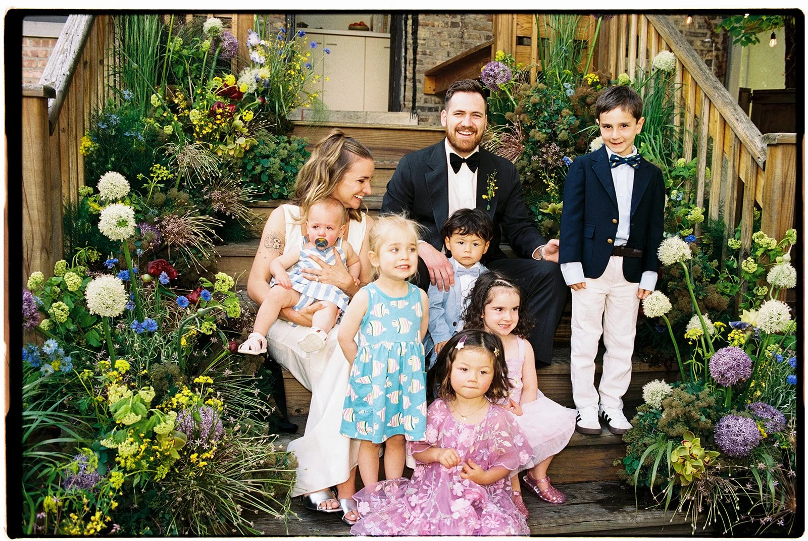 A family portrait on wooden steps surrounded by colorful flowers. An adult woman with a baby, a man in a black tuxedo, and several children of various ages wearing formal and casual clothes are smiling for the camera in an outdoor setting.
