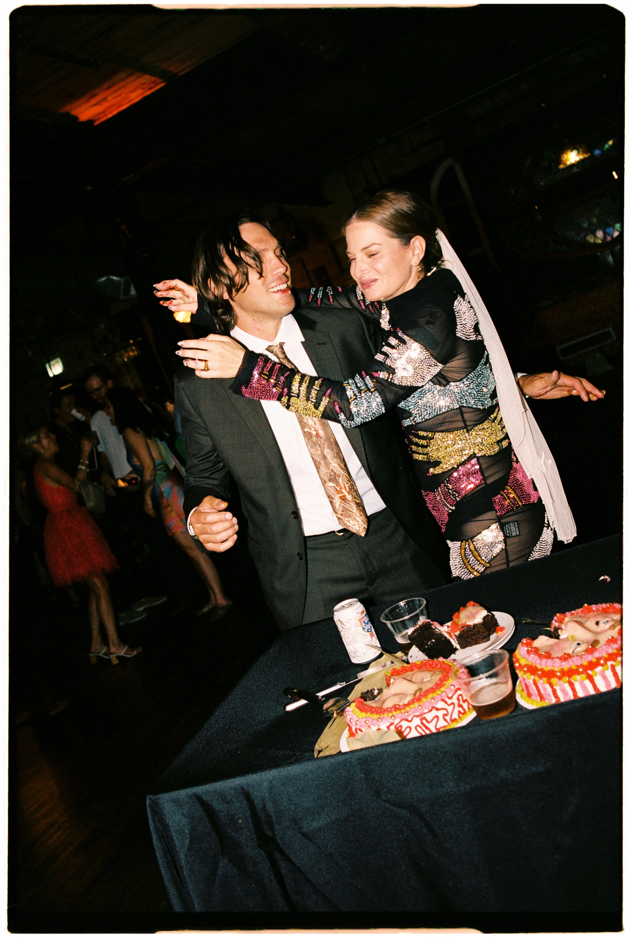 A woman and a man are dancing together at a party or celebration. The woman is wearing a colorful, patterned dress and has a veil. The man is wearing a dark suit with a patterned tie. In the foreground, there are birthday cakes with colorful decorati