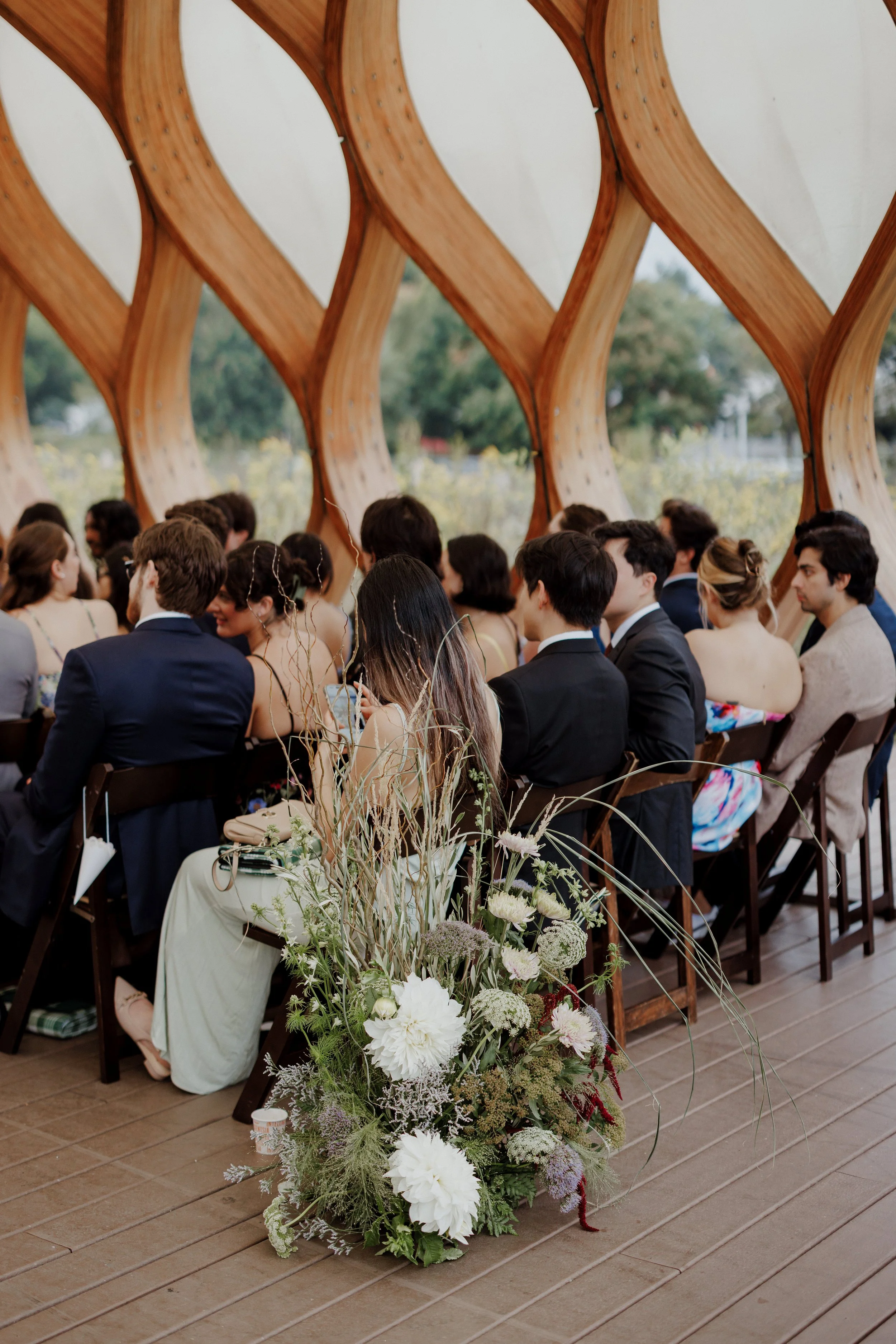 A diverse group of people dressed in formal attire sitting in rows at an outdoor wedding ceremony, with a large floral arrangement in the foreground and wooden architectural design in the background.