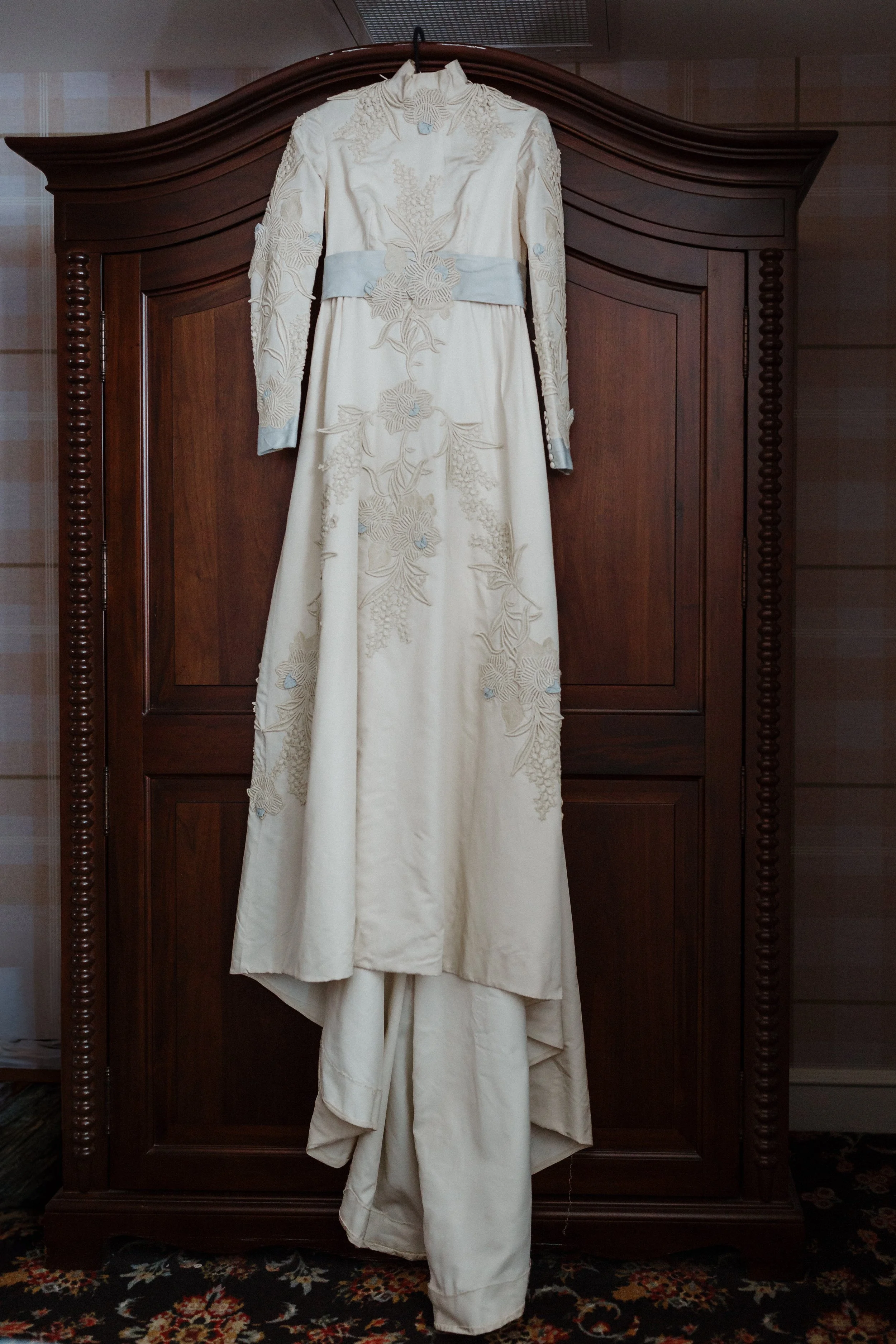 A white vintage wedding dress with floral embroidery hanging on a wooden wardrobe.