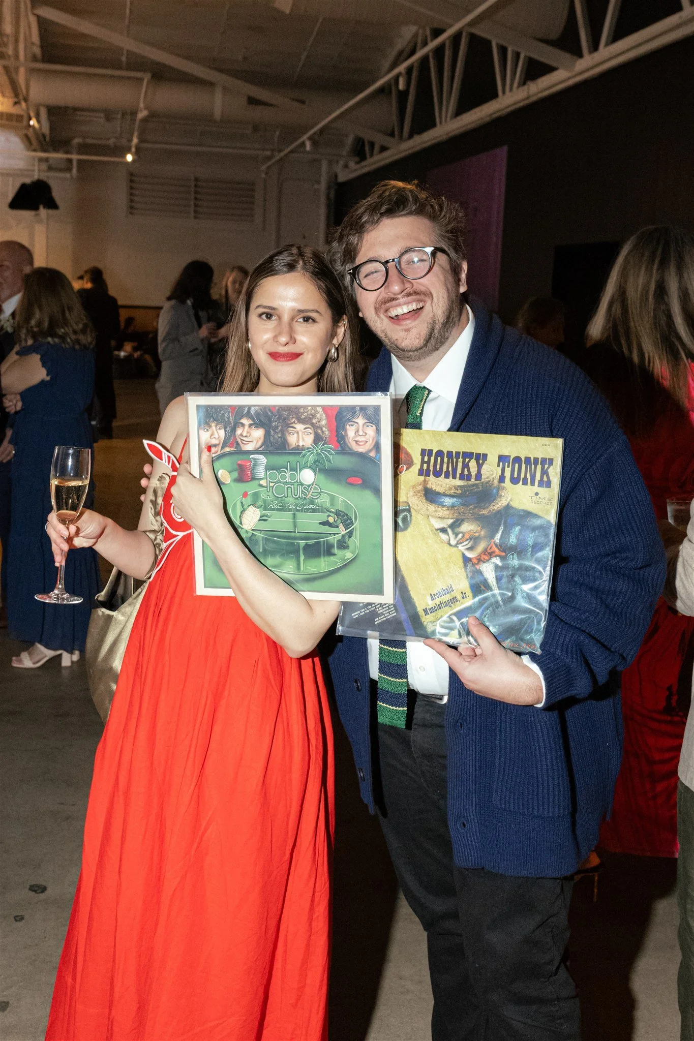 A man and woman at a social event holding vinyl records and a glass of champagne. The woman is wearing a red dress and the man is wearing glasses, a suit, and a sweater. They are smiling and standing close together.