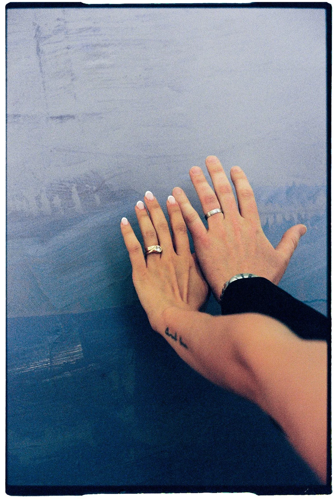 Two hands with wedding rings and a tattoo, resting on a glass surface.