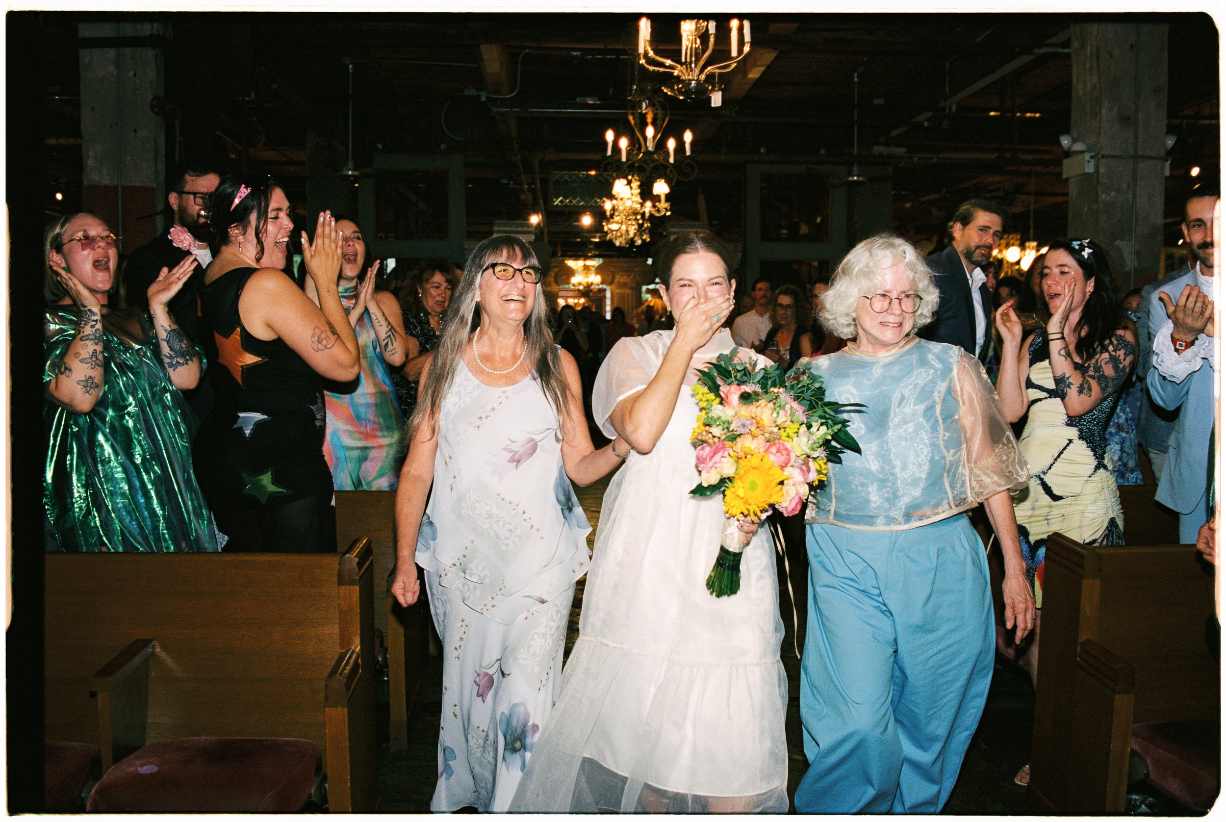 Group of women and men celebrating at a wedding, including an older woman with blonde curly hair and glasses, a bride with a bouquet of flowers, and guests joyfully interacting in a warmly lit venue with chandeliers.