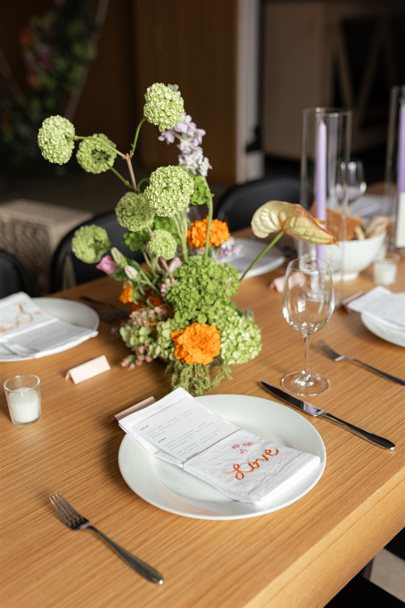 A table setting with a white plate, silverware, a napkin embroidered with the word 'Love', a menu, a wine glass, a small candle, and a tall floral centerpiece with green, purple, and orange flowers.