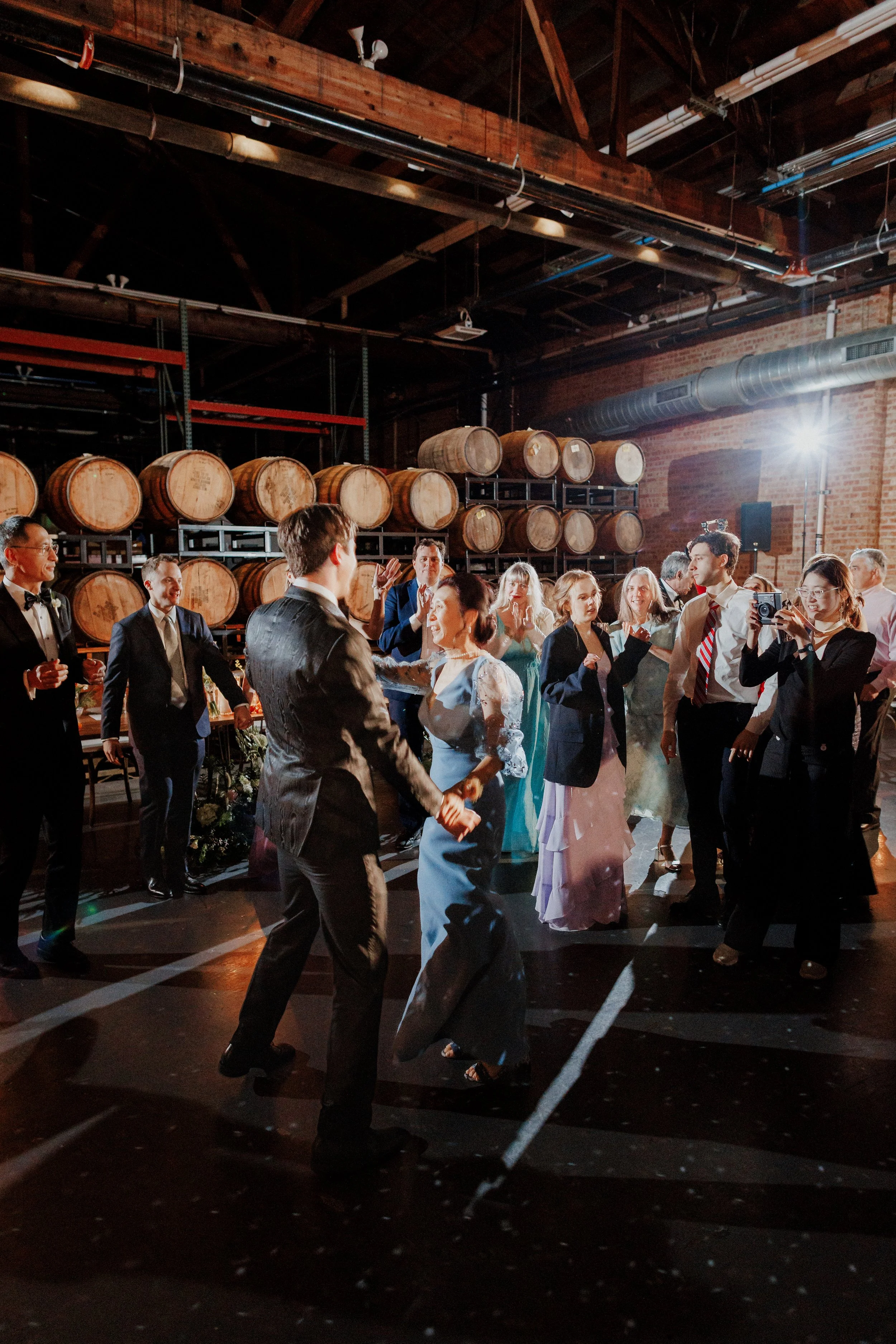 People dancing and celebrating at a wedding reception in a brewery with brick walls and stacked barrels in the background.