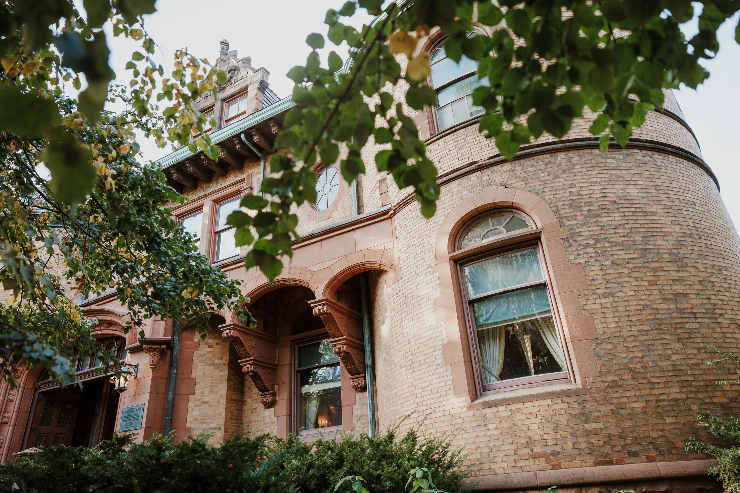 A historic brick building with tall arched windows and ornate architectural details, partially obscured by green leafy tree branches in the foreground.