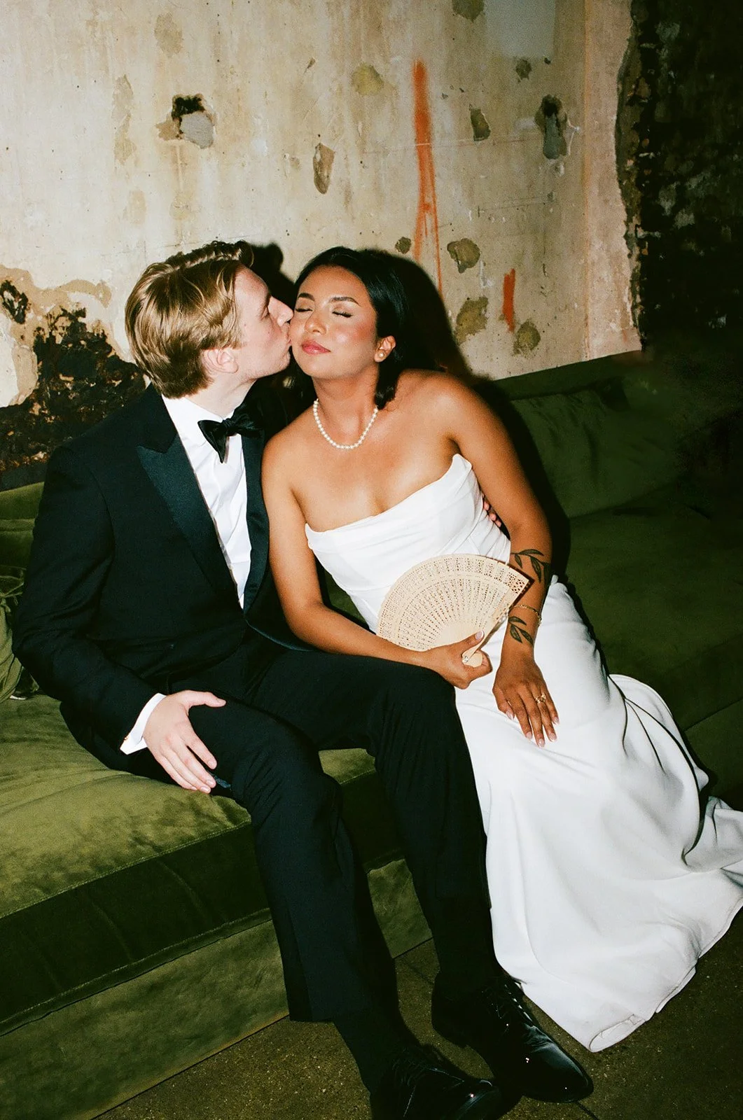 A man in a tuxedo is kissing a woman in a white strapless gown on the cheek. The woman has dark hair, is wearing a pearl necklace, and is holding a fan. They are sitting on a green sofa against a rustic, peeling wall.