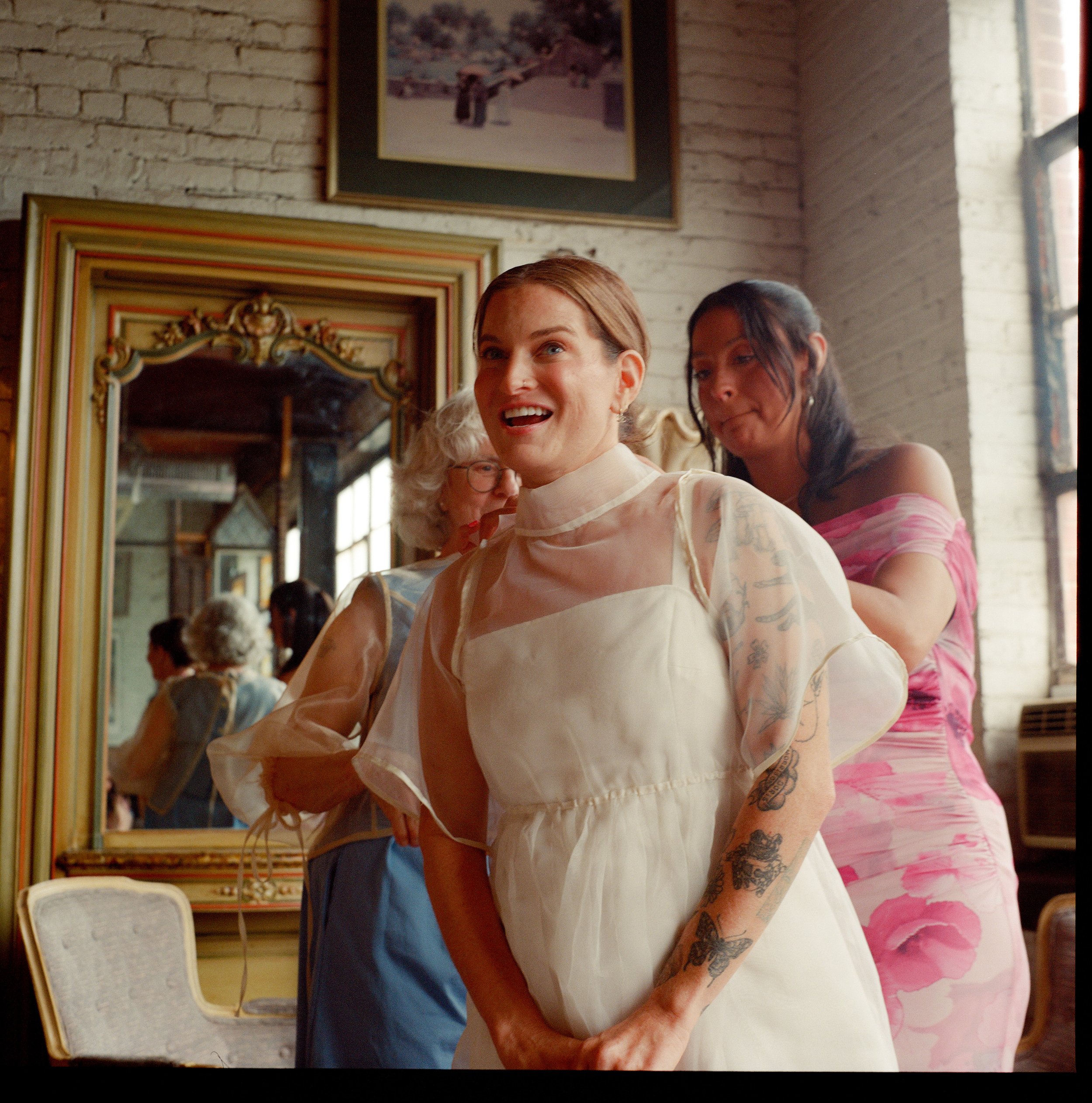 Two women smiling and having fun together, one wearing a white dress with tattoos on her arm, the other in a pink and white striped dress, in a room with a vintage mirror and framed artwork on the wall.
