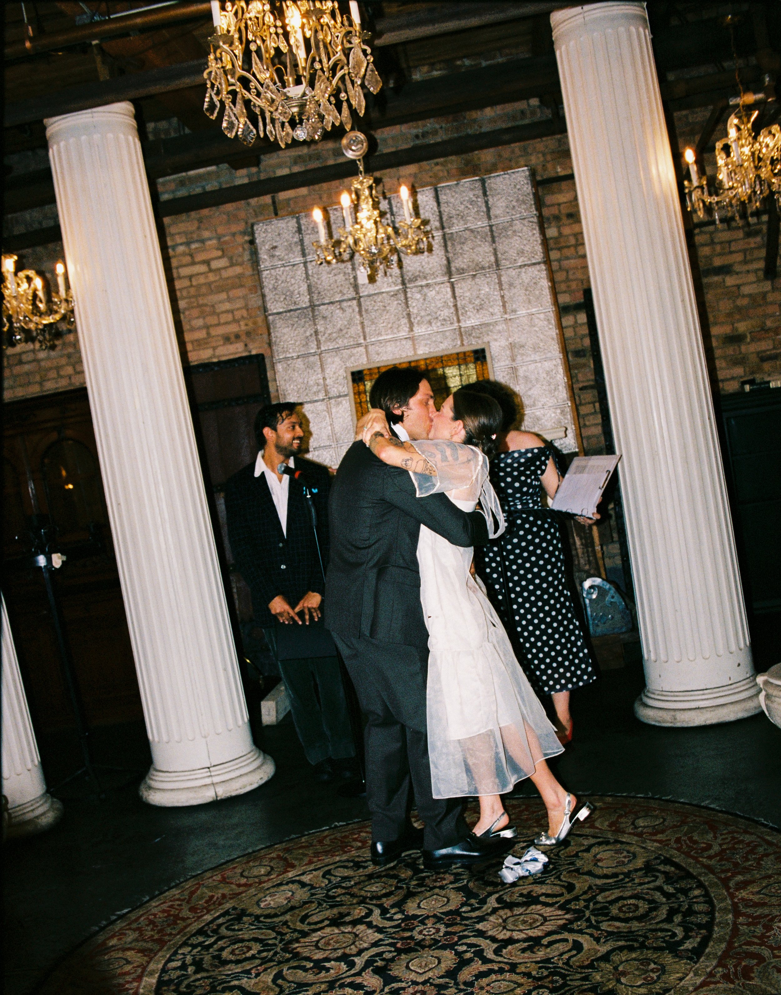A couple is kissing at a wedding reception, standing on a patterned rug under chandeliers, with a brick wall background and white columns nearby. The bride is in a white dress and silver heels, and the groom is in a dark suit.