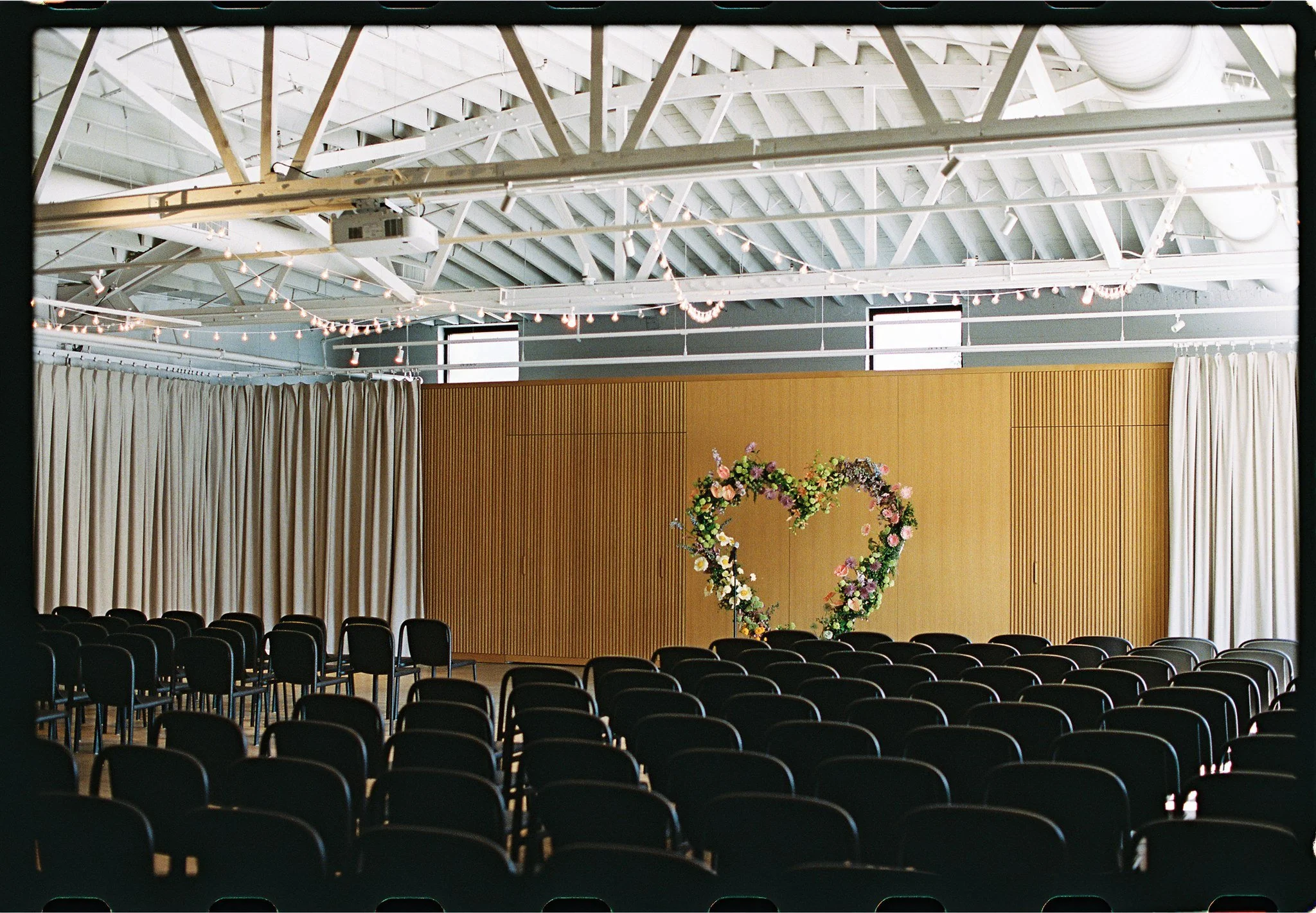 An indoor event space with rows of black chairs facing a stage decorated with a floral heart arrangement. The stage has a wooden backdrop, cream drapes on the sides, and string lights hanging from the ceiling. The ceiling has exposed white beams and 