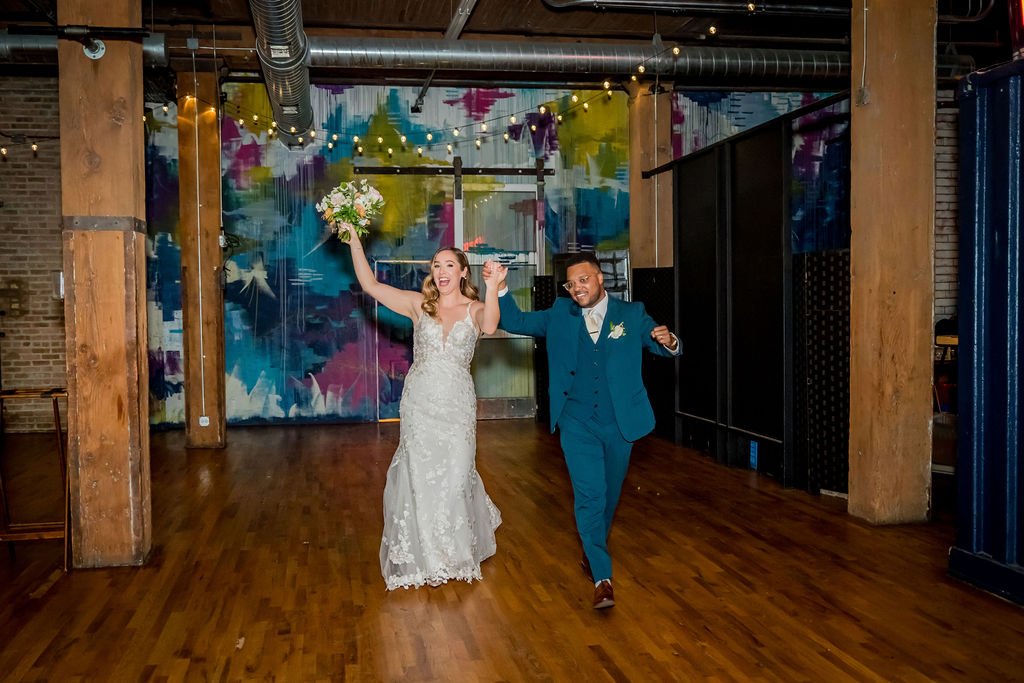 A bride in a white wedding dress holding a bouquet, and a groom in a blue suit dancing together in a decorated venue with string lights and colorful wall art.