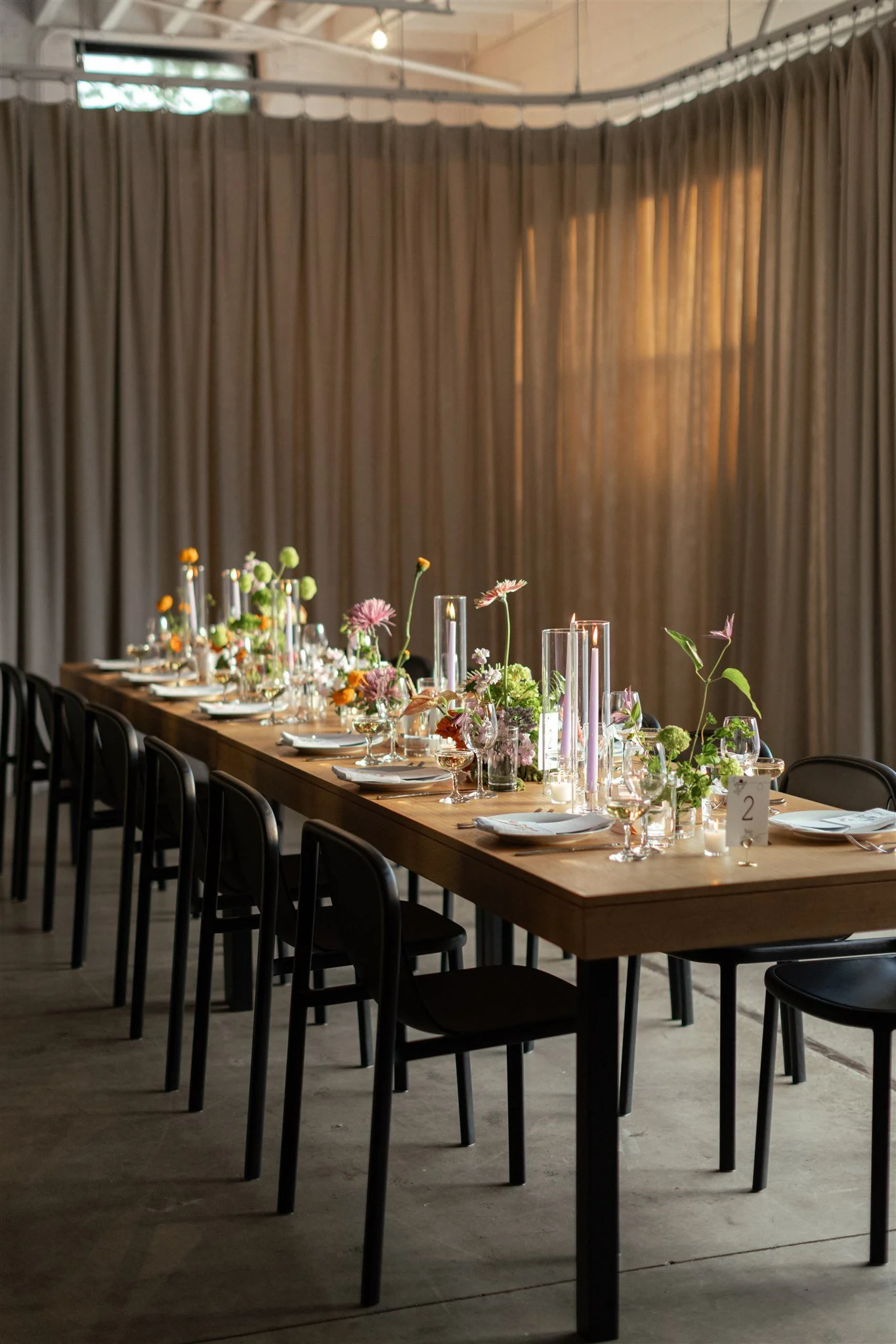 A long wooden dining table decorated with flowers and candles, set for a formal event, with black chairs around it, and a beige curtain backdrop.