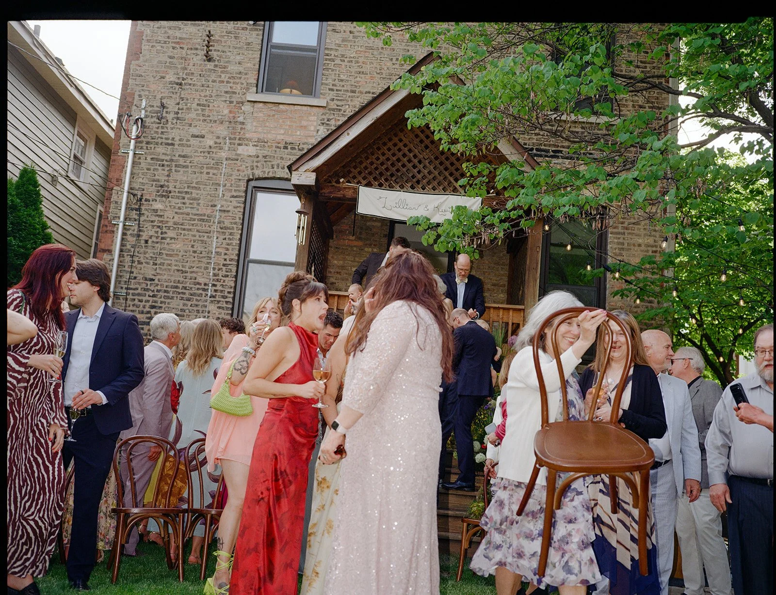 People socializing at an outdoor wedding reception in a backyard with brick building walls and trees, some holding drinks, under a wooden canopy.
