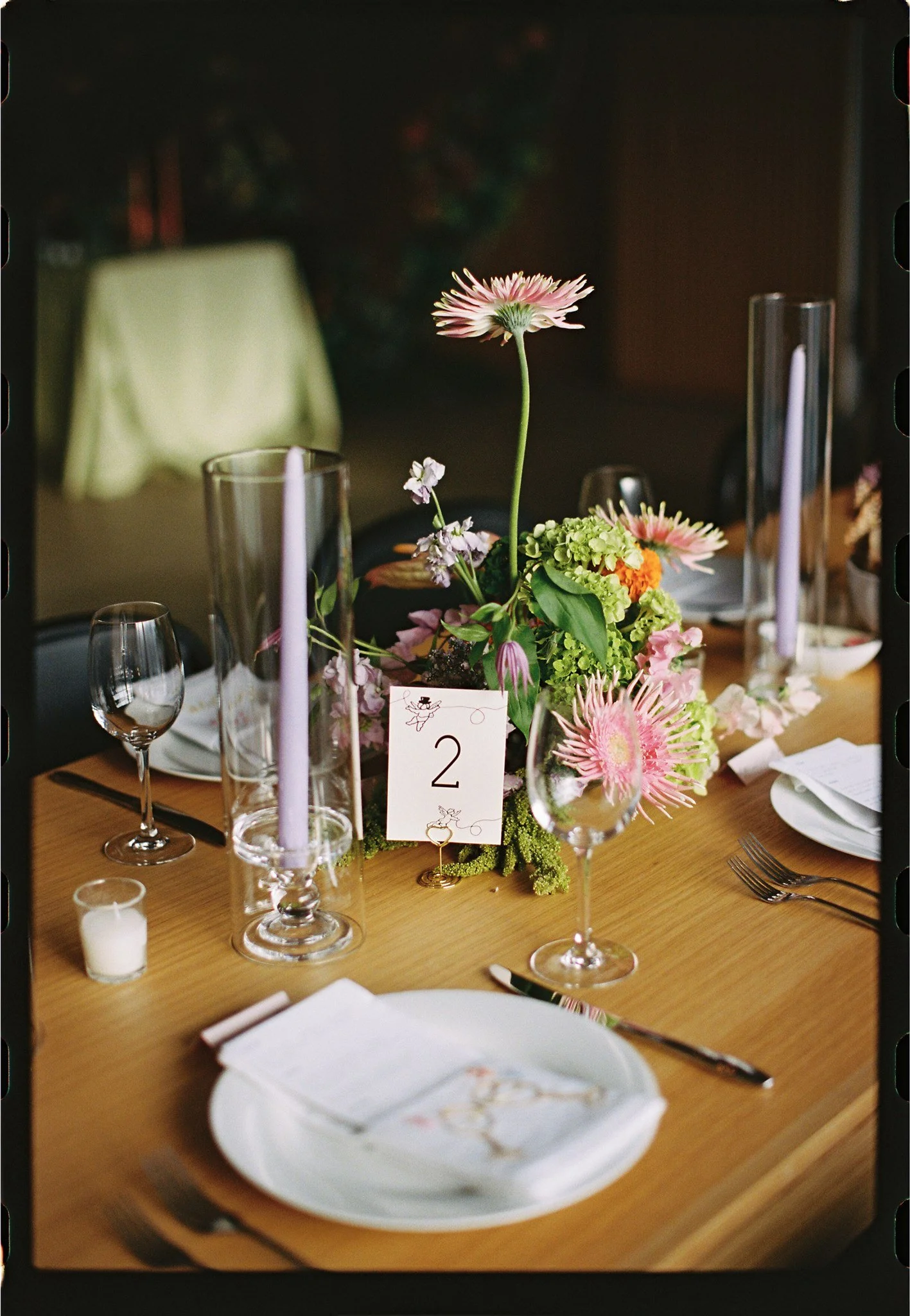 Table setting at a formal event with a floral centerpiece featuring pink and green flowers, wine glasses, white plates, a napkin, a small candle, and a table number card with the number 2.
