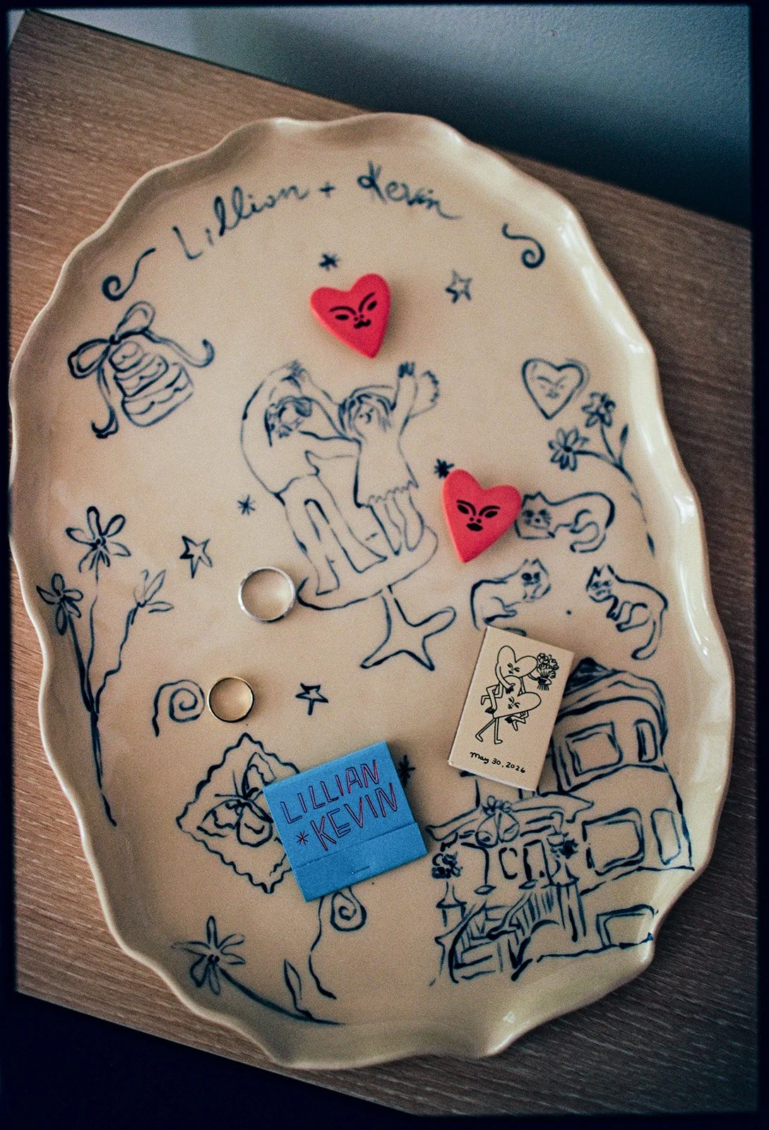 Decorative ceramic tray with hand-drawn illustrations of children, animals, and flowers, featuring two pink heart-shaped items with angry face designs, a small illustration of a girl with a heart, a blue sticky note with handwritten names 'Lillian an
