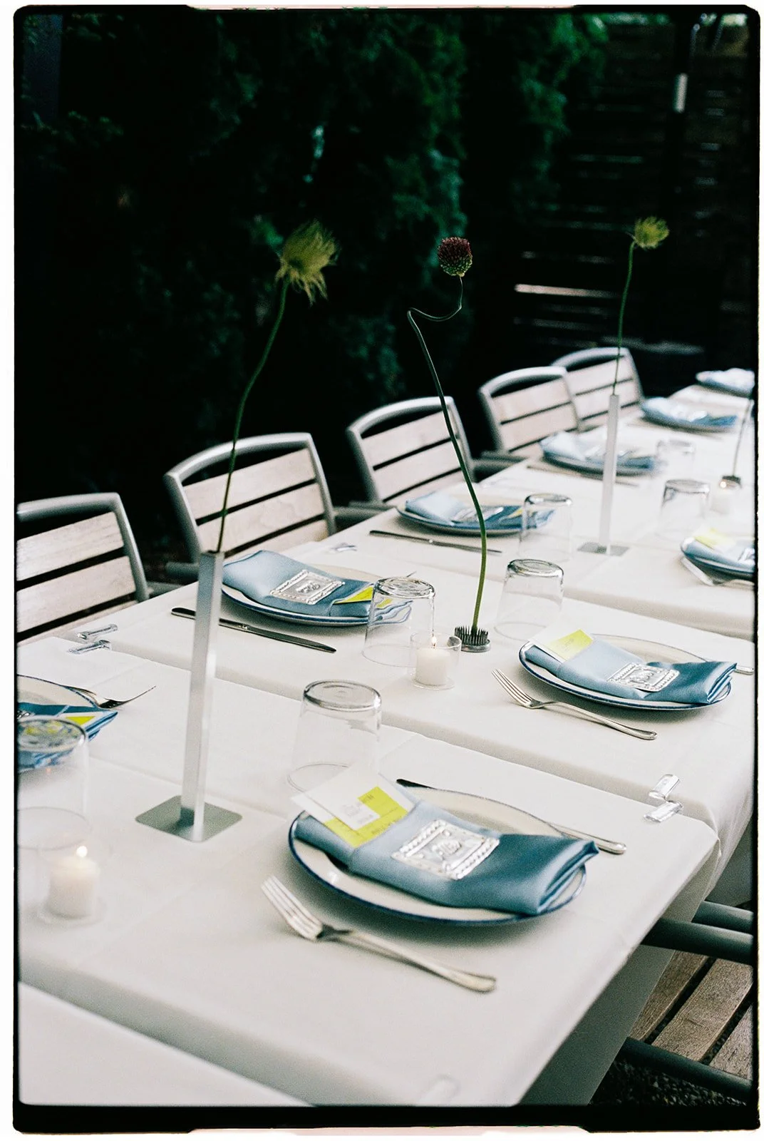 A long dining table set for an event with white tablecloths, blue napkins, and place settings including glasses, plates, and silverware. Tall, slender floral centerpieces with minimal flowers and candle decorations are arranged along the table. Woode