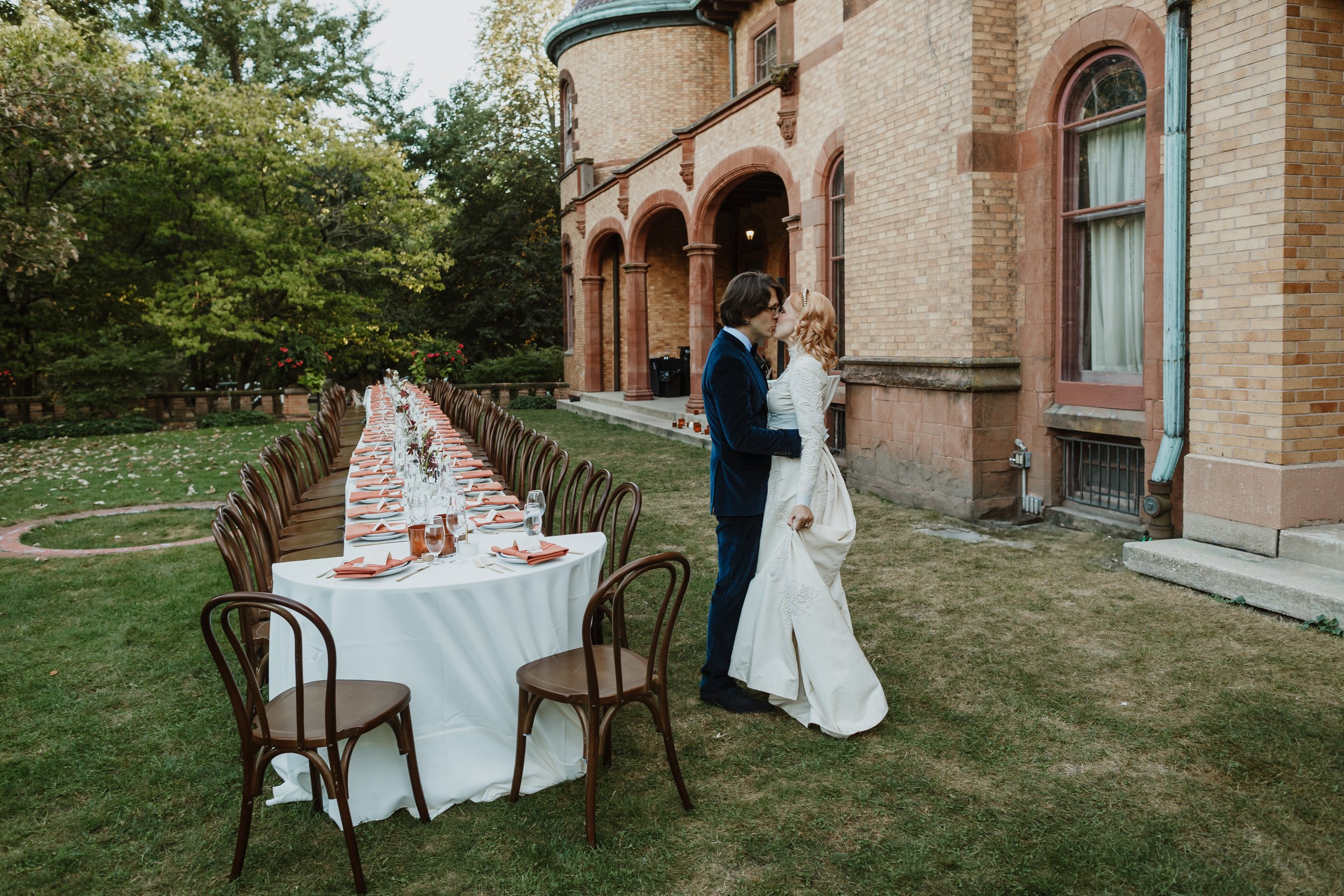 A couple is kissing outdoors during a wedding reception at a long tablesetting on the lawn in front of a large brick house with arched windows and stone accents. The tables are decorated with white tablecloths, glasses, and napkins, and surrounded by