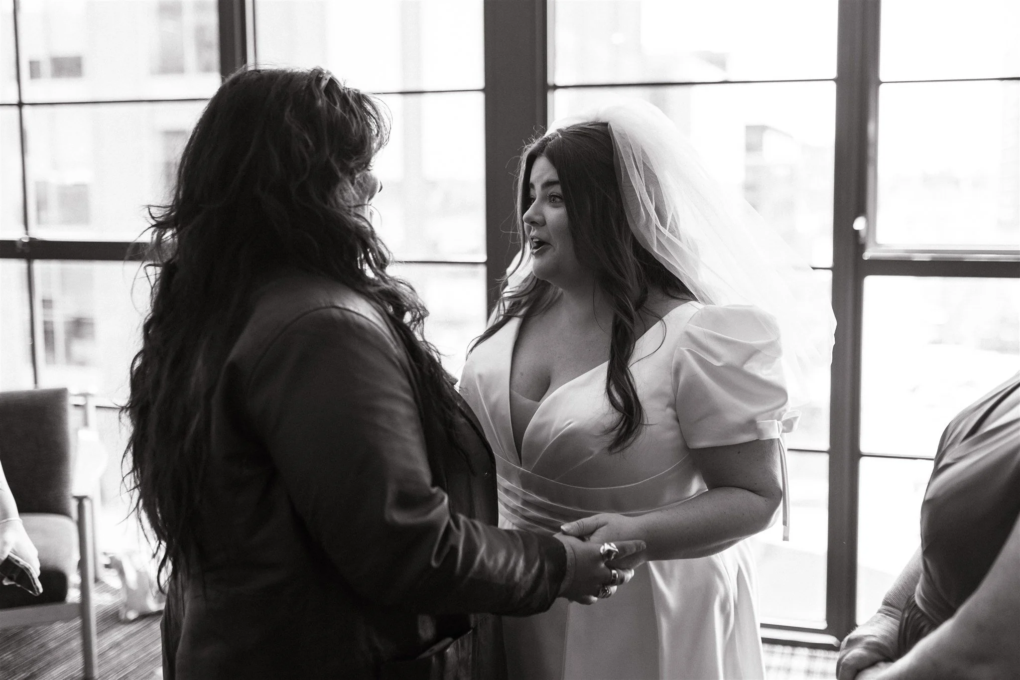 A black and white photo of a woman in a white dress and veil holding hands with a woman in a dark jacket, standing close together in front of large windows.