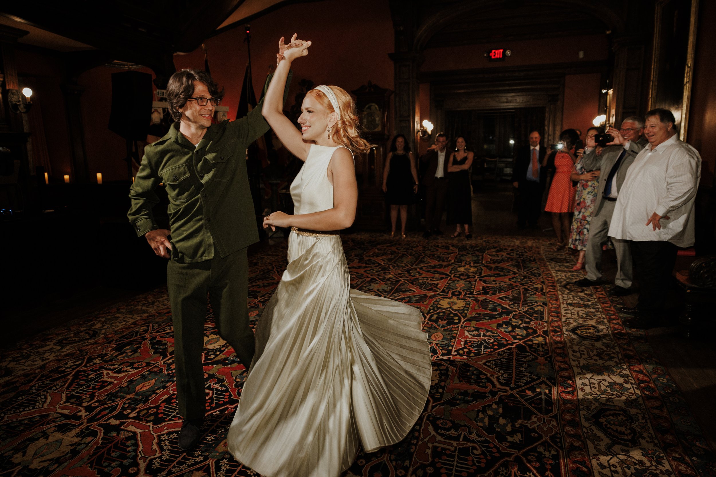 A bride and groom dancing at their wedding reception with guests watching and taking photos in a dimly lit, decorated banquet hall.