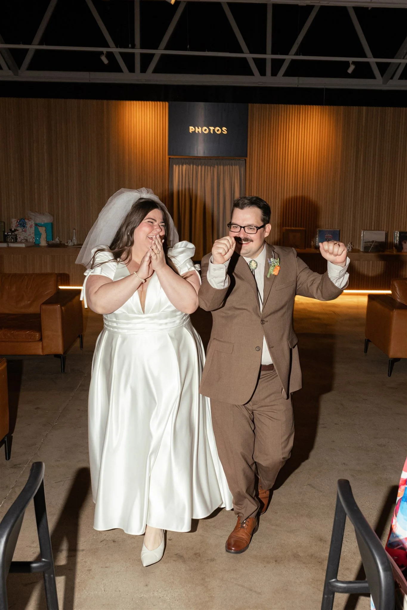 A bride and groom celebrating at their wedding reception, with the bride wearing a white wedding dress and the groom in a brown suit, both smiling and showing excitement.