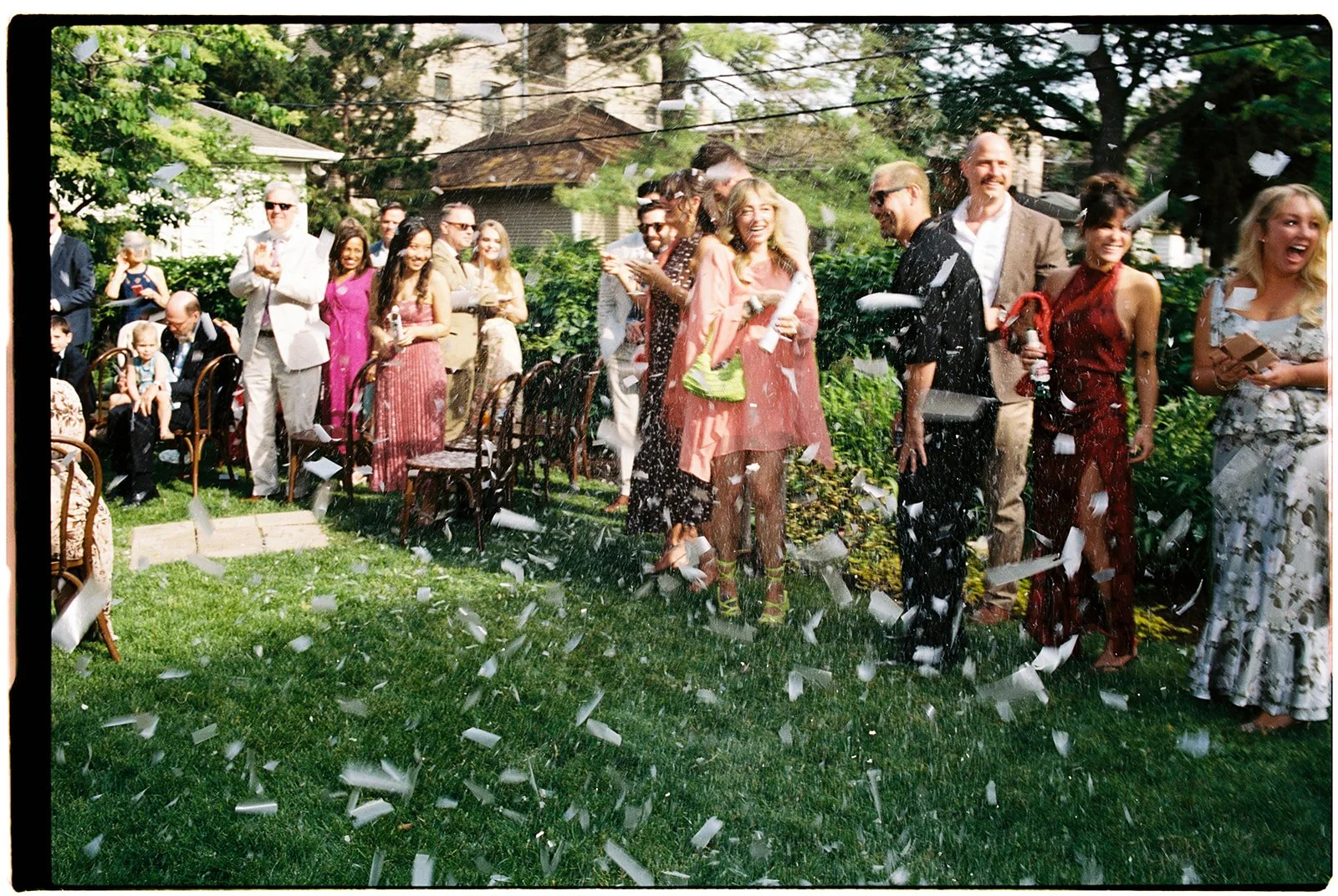 People celebrating outdoors with confetti on a green lawn, some laughing and holding drinks, in daytime with trees and houses in background.