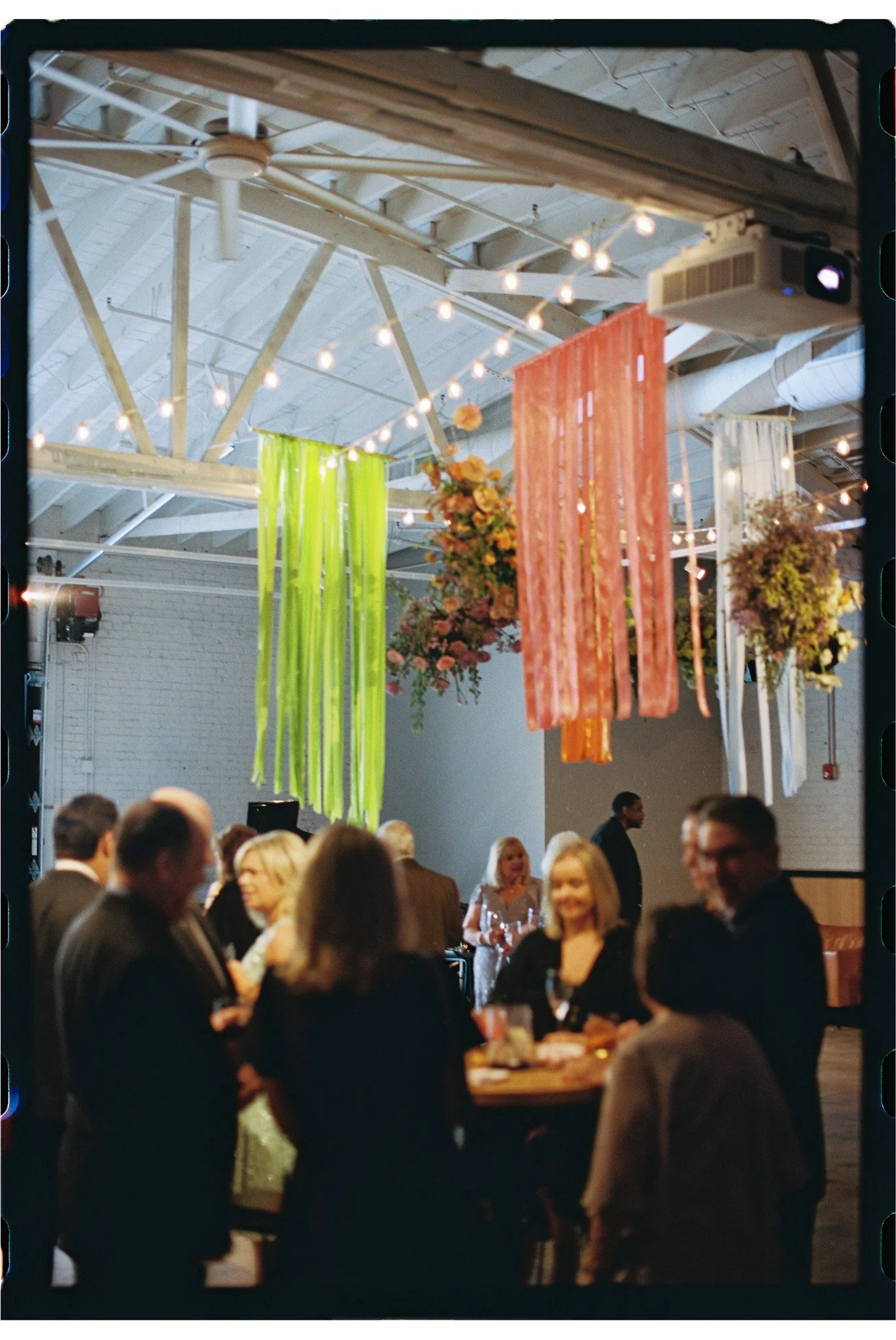 A crowded social gathering indoors with hanging string lights, colorful streamers, and floral decorations, with people dressed in formal attire engaging in conversation.