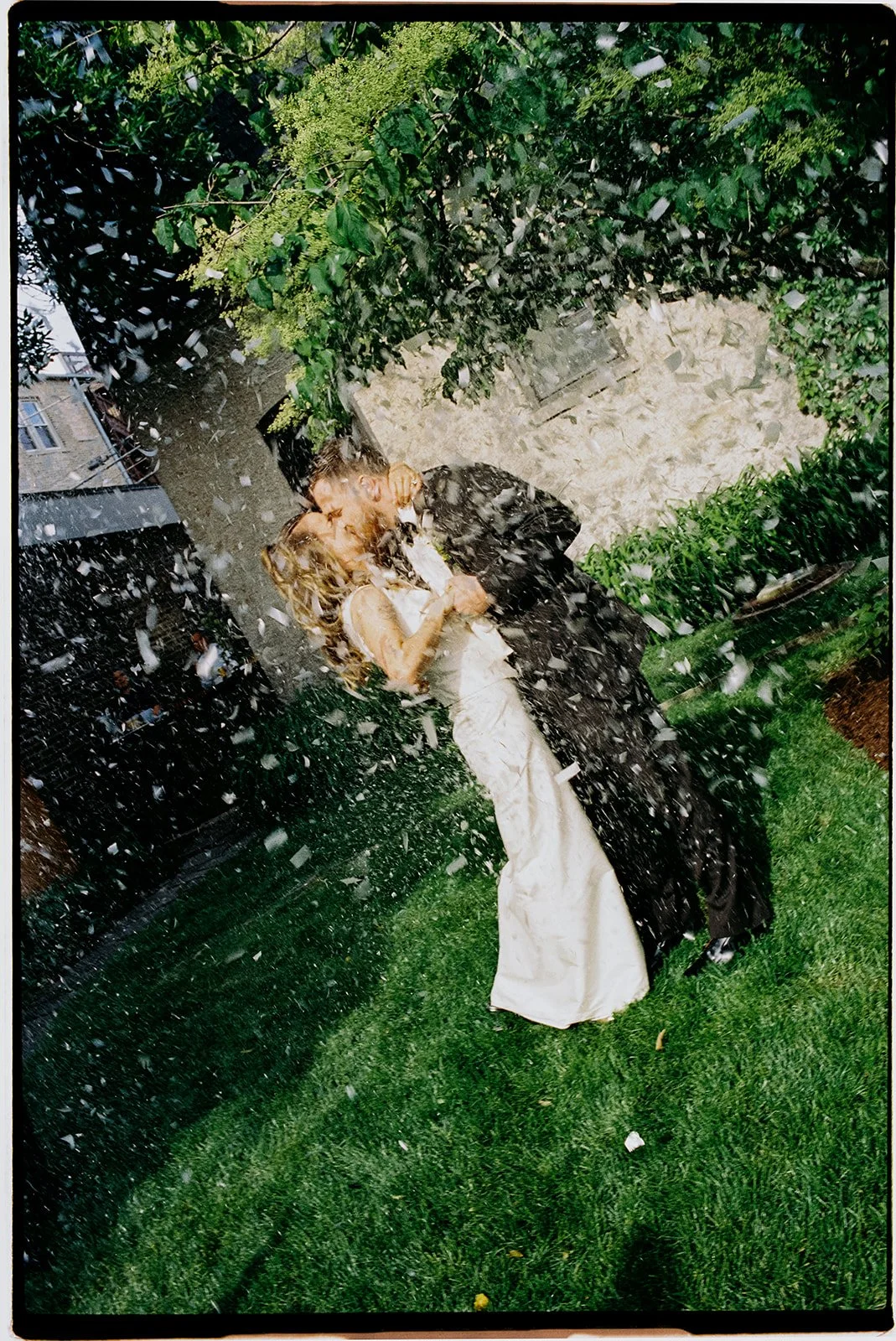 A couple wearing wedding attire kissing under a garden sprinkler on a lawn during the daytime.