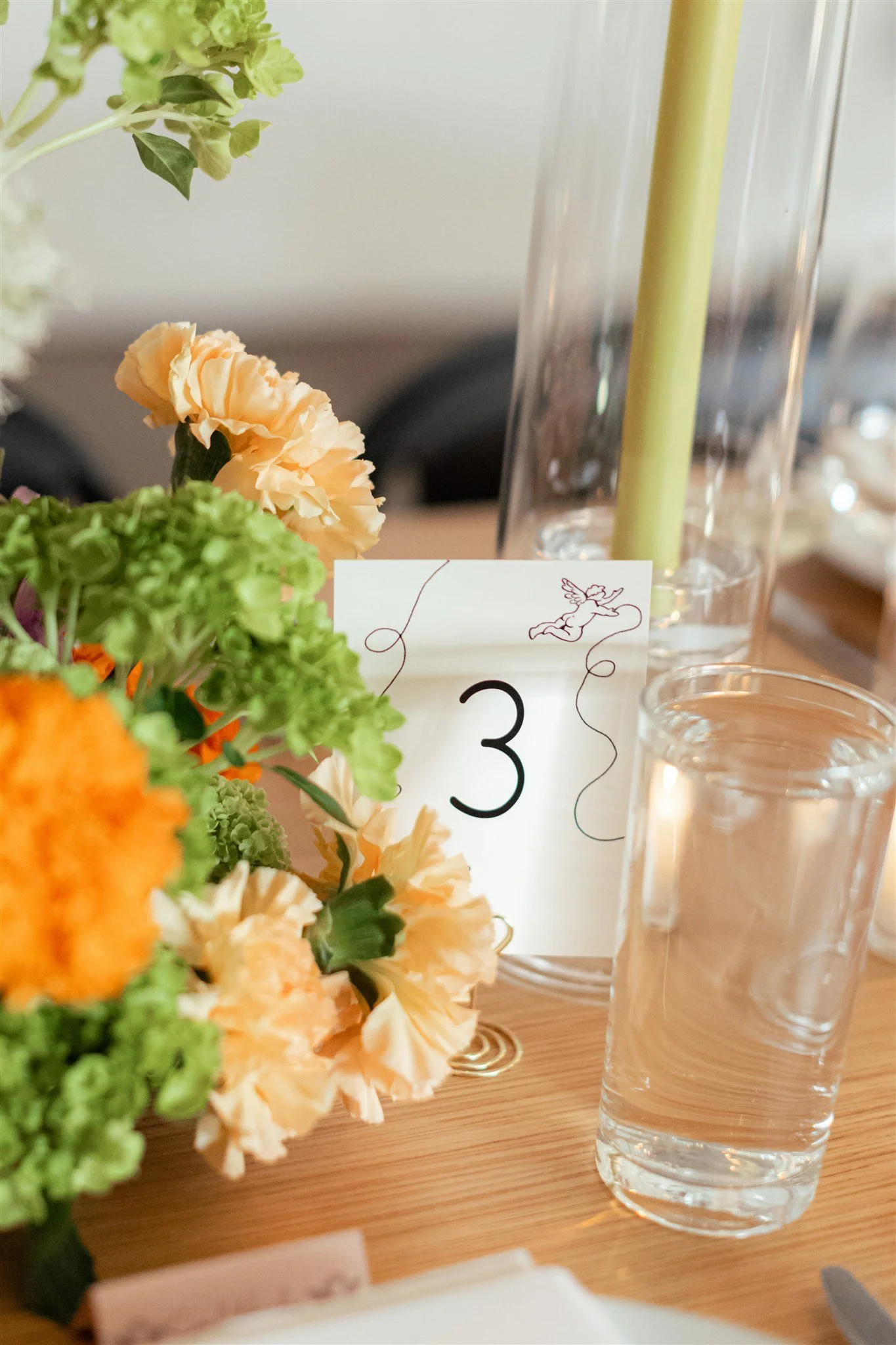 A table with a floral centerpiece, a small card with a number 3, a tall clear glass vase, and empty clear glasses on a wooden surface.
