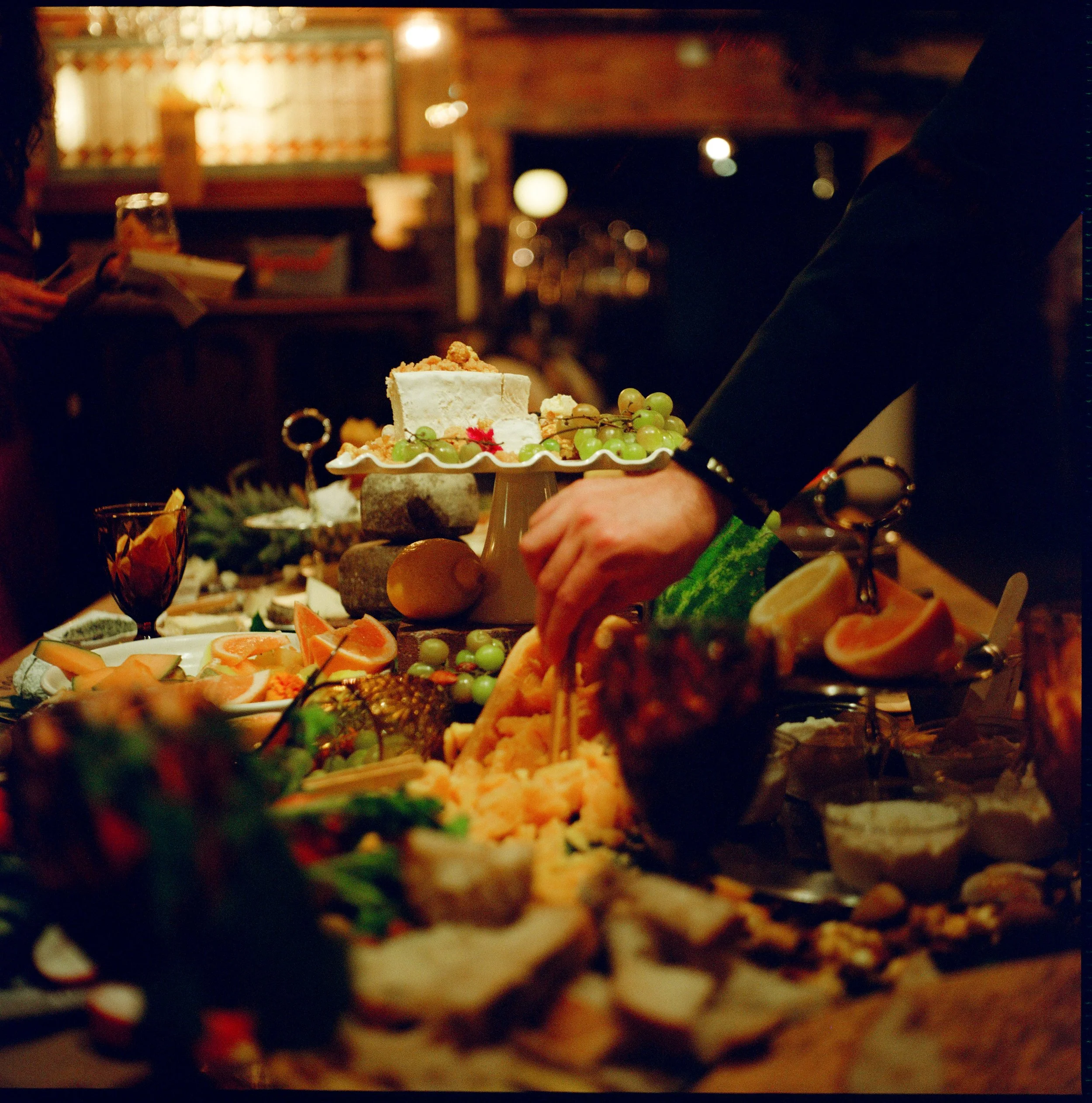 A blurred image of a person serving or presenting a dessert cake decorated with grapes on a table. The background appears to be a cozy indoor setting with warm lighting.