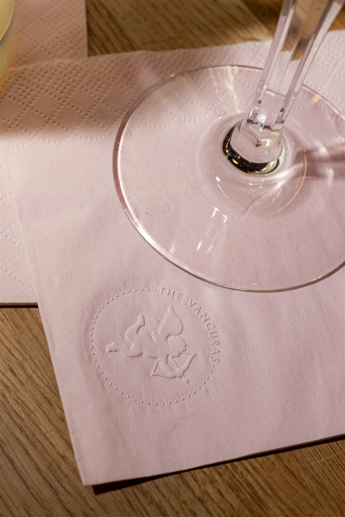 Close-up of a pink paper napkin with an embossed logo of a unicorn and the word 'VANQUERAS,' placed on a wooden table with a glass of water.