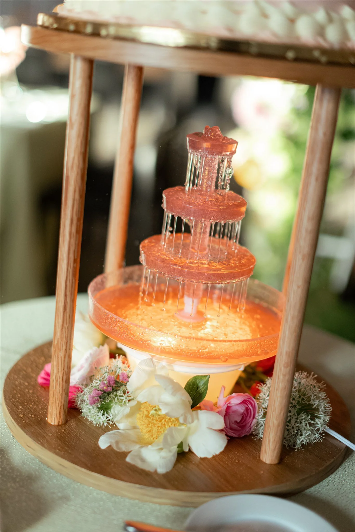 A tiered orange-colored water fountain with water flowing from each tier. The fountain is surrounded by flowers and placed on a wooden stand with four wooden legs.