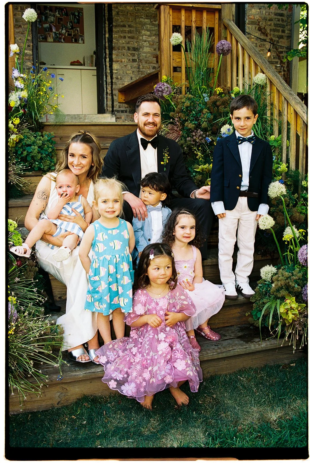 A group of people, including adults and children, dressed in formal attire, sitting and standing on wooden stairs outdoors surrounded by colorful flowers and greenery, with a brick building in the background.