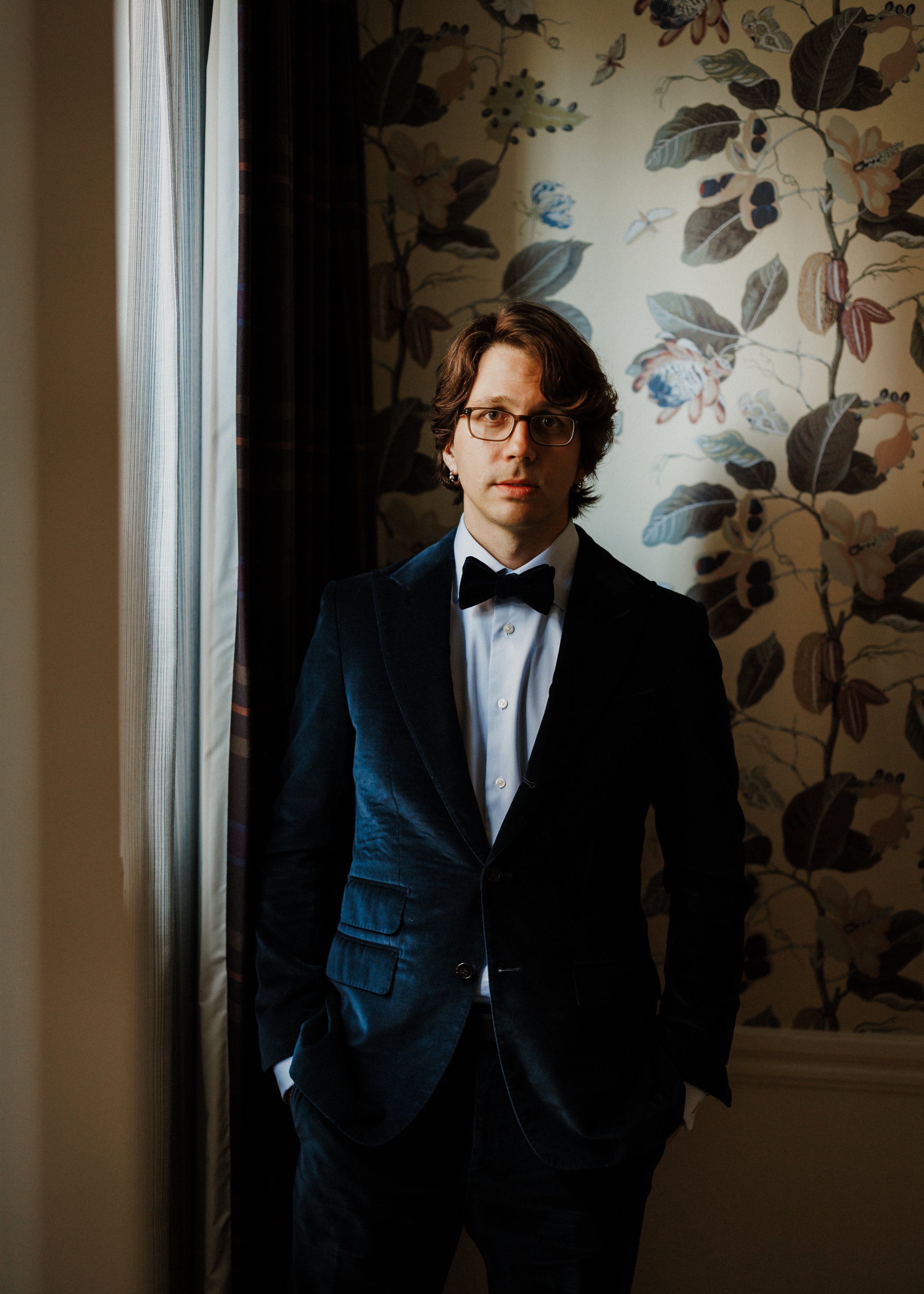 A man in a tuxedo and glasses standing indoors near a floral wallpapered wall and a window with curtains, looking at the camera.