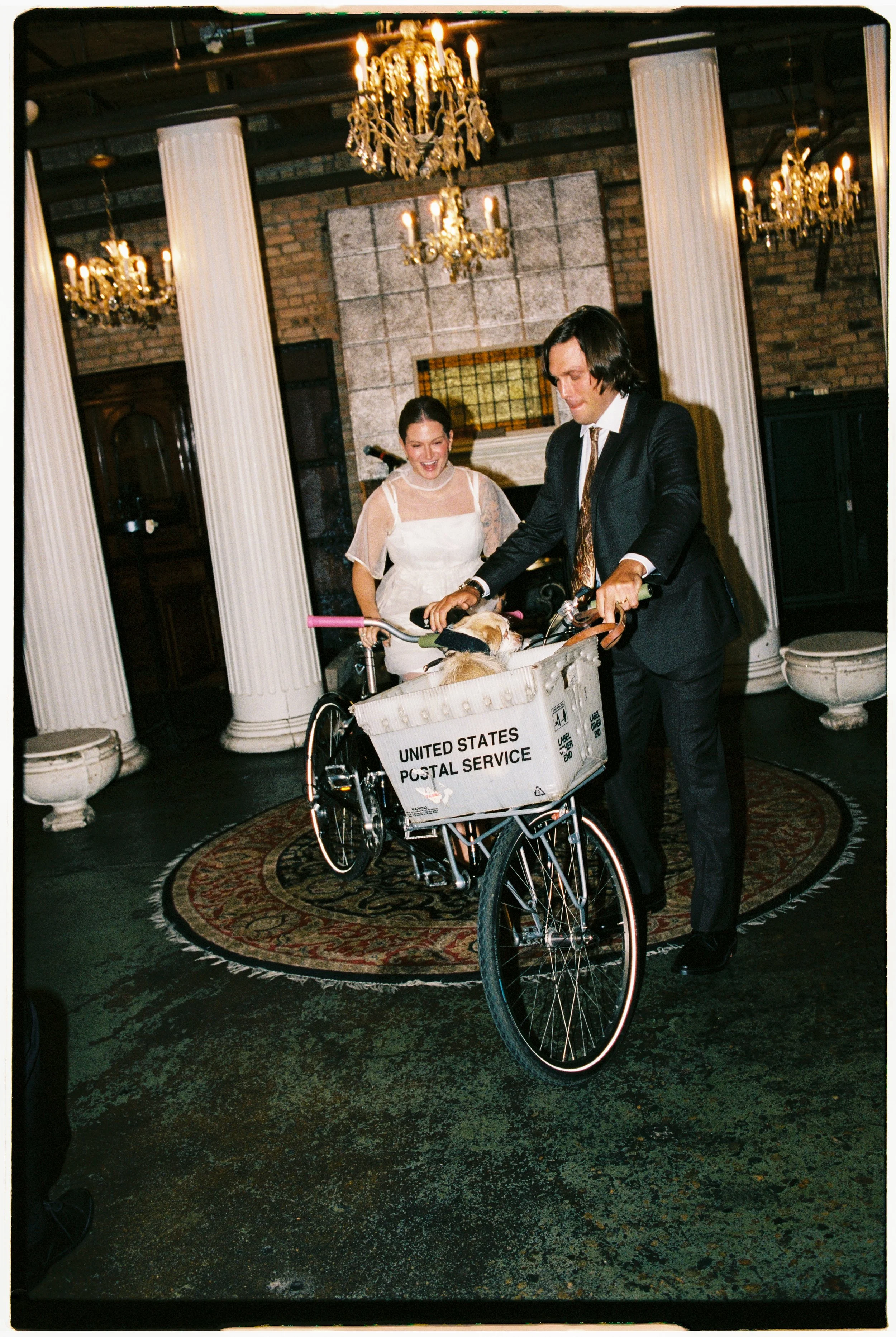 A man in a suit with long dark hair is riding a bicycle with a canine in the basket, while a woman in a white dress watches and smiles in a room with ornate chandeliers, white columns, and brick walls.