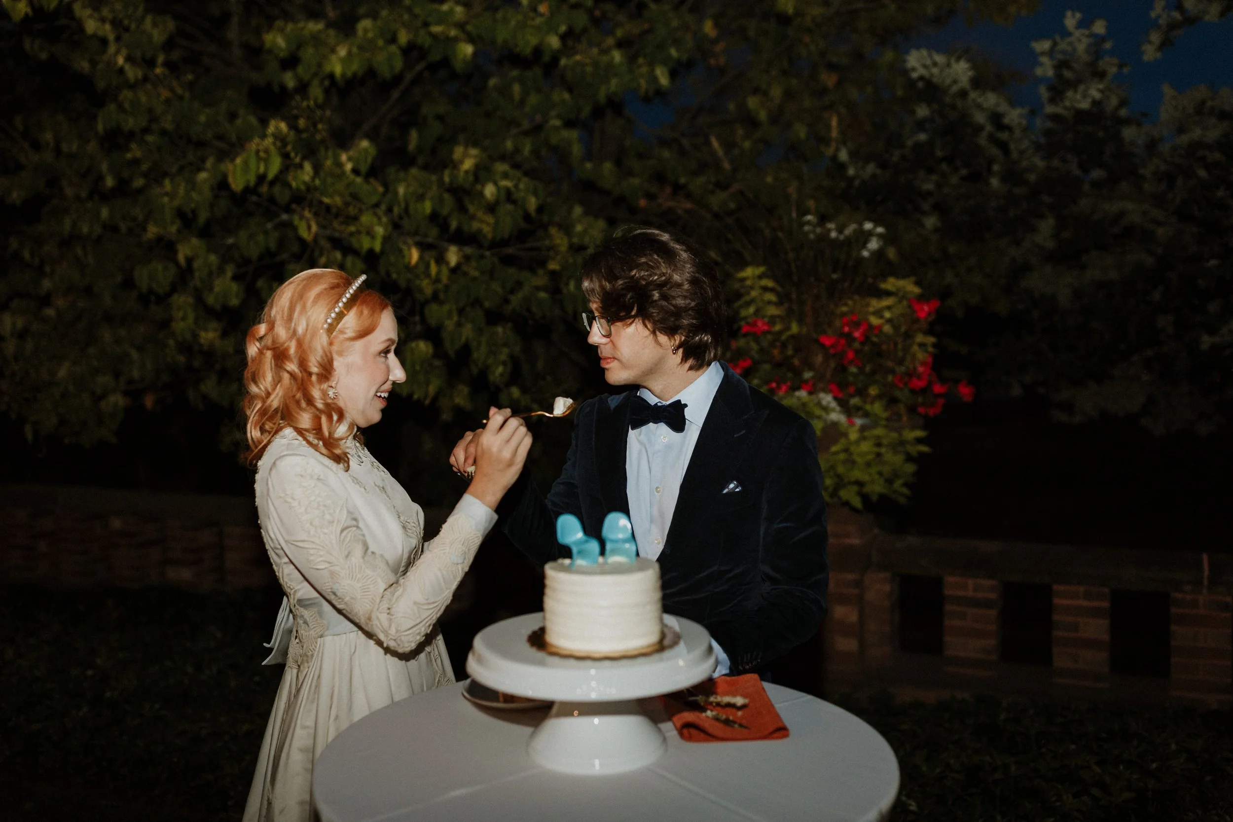 A woman with red hair in a white dress feeds a man with dark hair and glasses with a fork at a celebration outdoors at night. A multi-tiered cake is on the table in front of them.