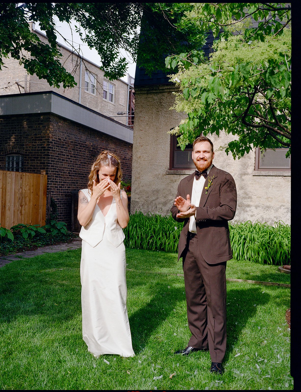 A bride and groom standing on a grassy lawn, celebrating outdoors under a tree. The bride is laughing and covering her face with her hands, wearing a white dress. The groom, dressed in a brown tuxedo with a black bow tie, is smiling and clapping, wit