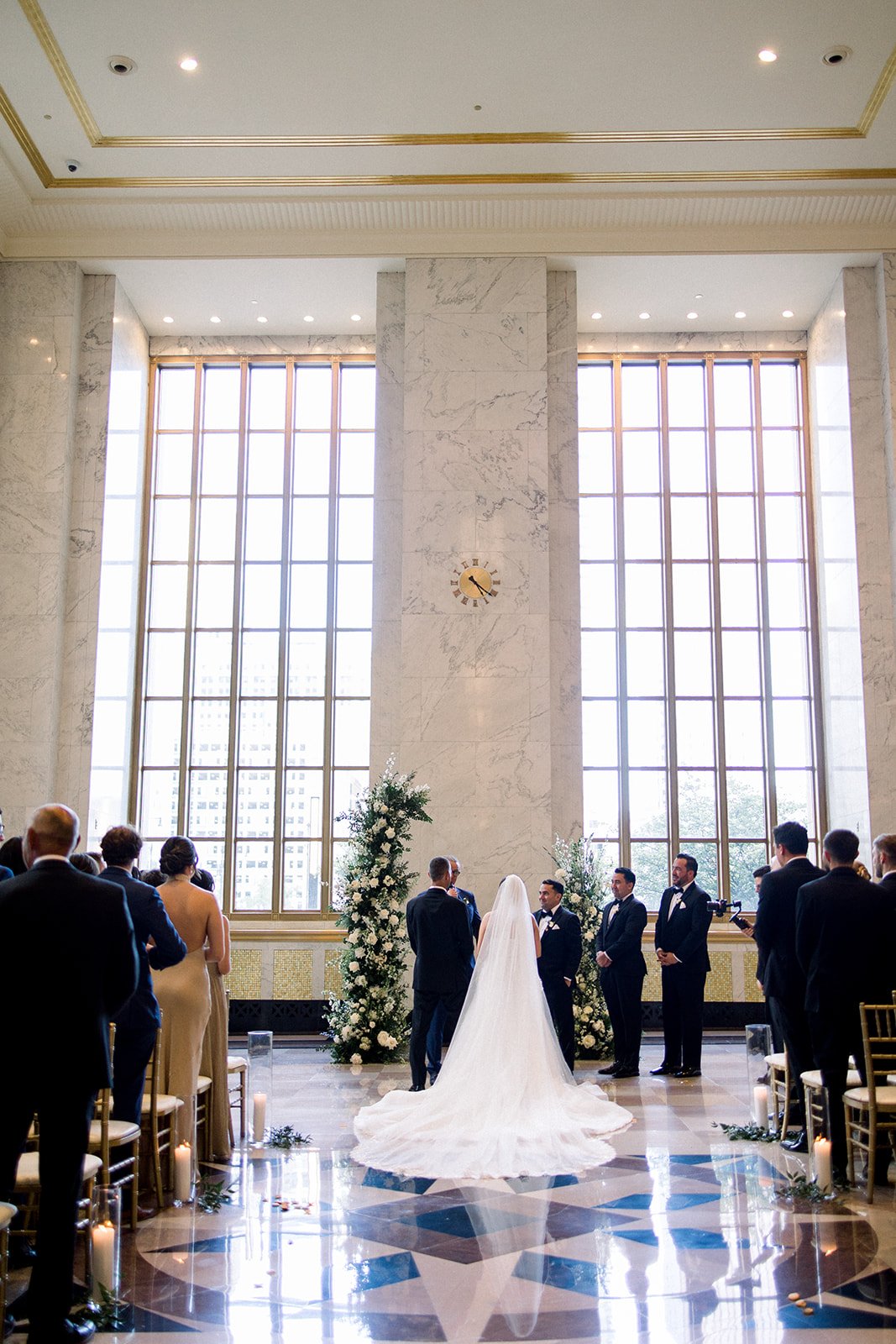 A wedding ceremony inside a grand hall with tall windows, floral arrangements, and a bride and groom standing at the altar with officiants and guests watching.