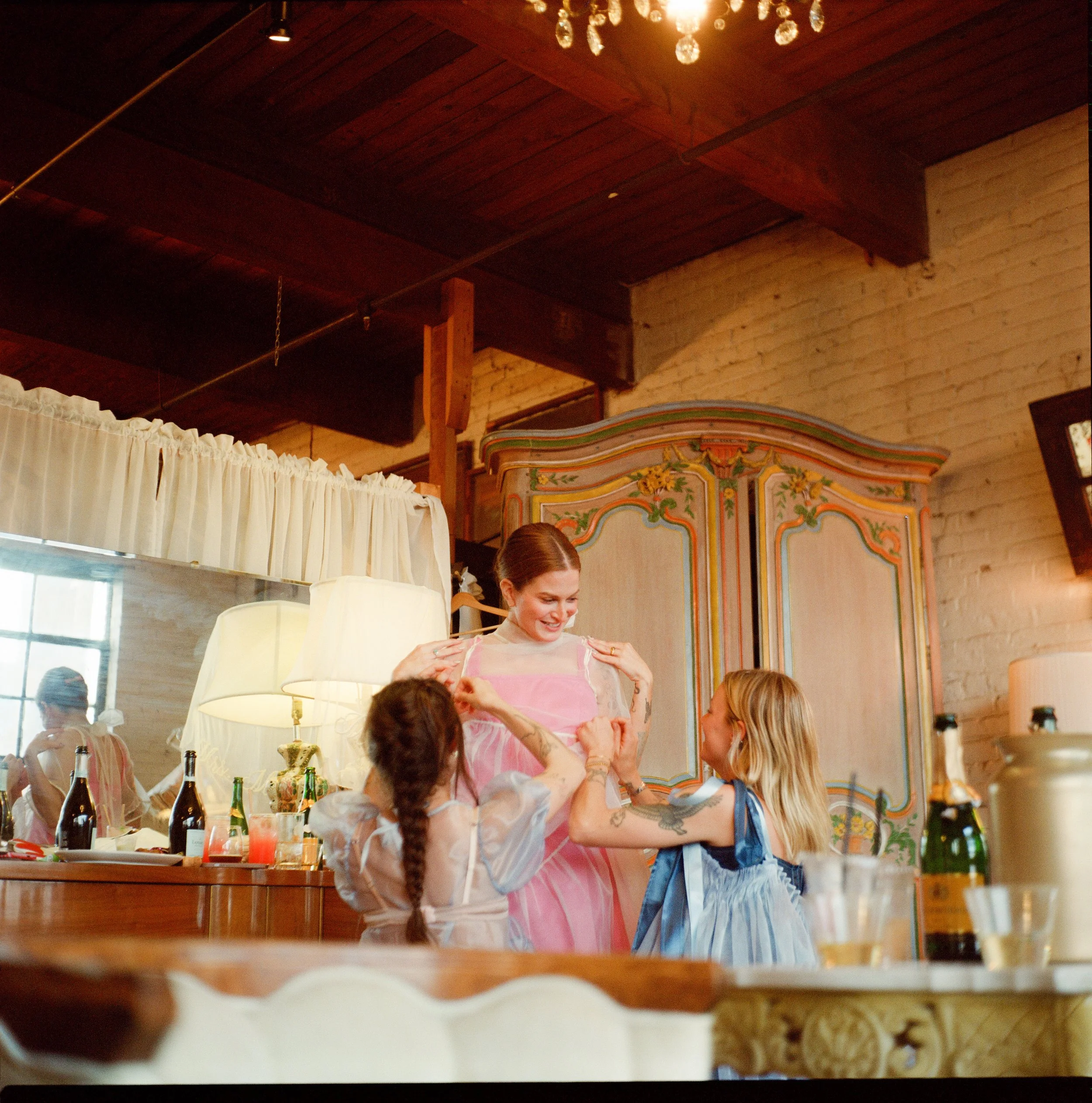 A woman in a pink and white striped shirt smiling and adjusting her top while a young girl with blonde hair in pigtails wearing a blue dress looks up at her. The scene takes place in a room with a rustic wooden ceiling, decorated to look like a carni