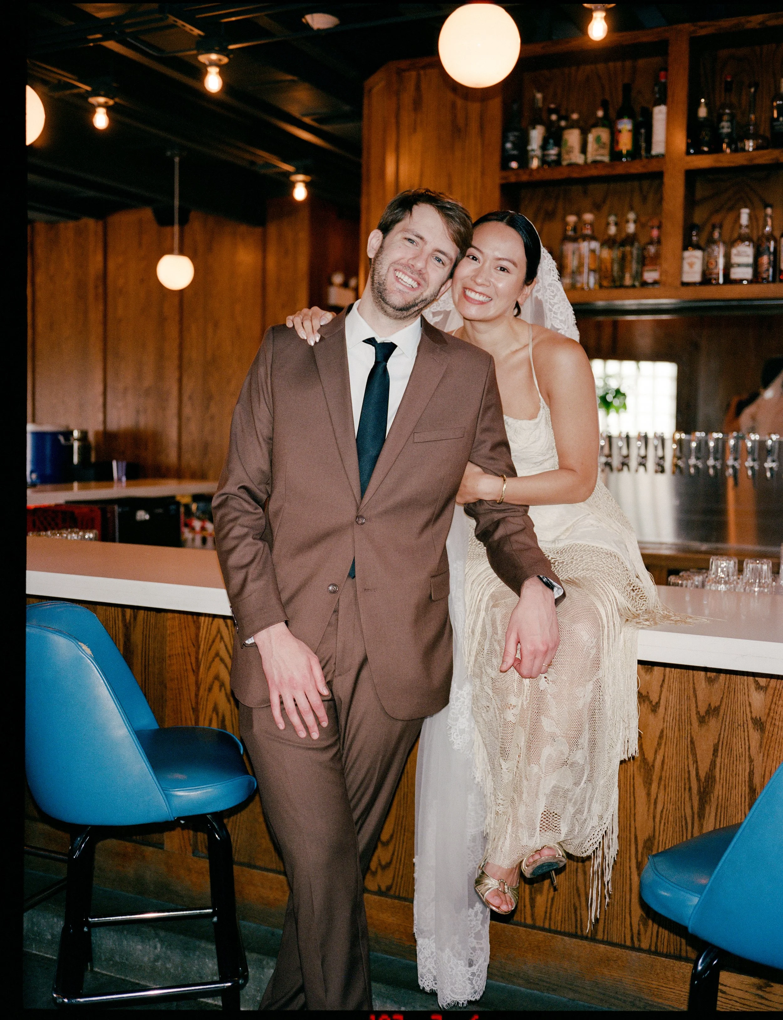 A man in a brown suit and a woman in a lace dress posing together in a bar or restaurant, smiling at the camera. The woman is wearing a lace dress with sandals, and the man is wearing a suit with a black tie. The background has wooden shelves with bottles and hanging light bulbs.