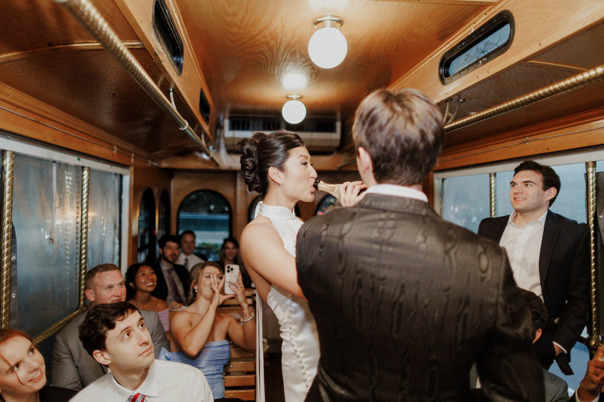 A group of wedding guests inside a vintage trolley bus, with a bride and groom taking a selfie together. The guests are smiling and capturing the moment on their phones.