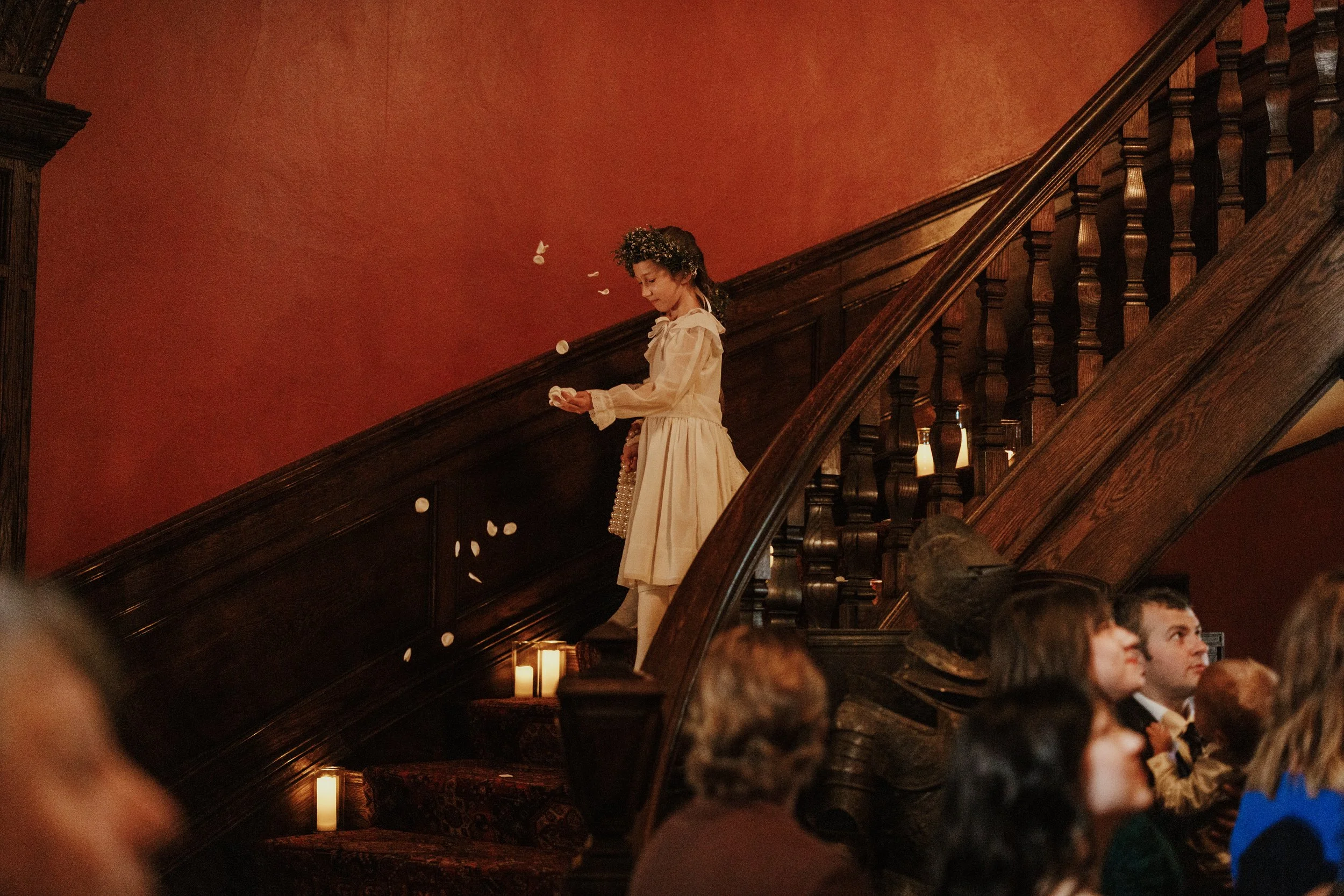 A girl wearing a beige dress and a flower crown is standing on a staircase tossing white flower petals, while several children sit on the floor below, watching.