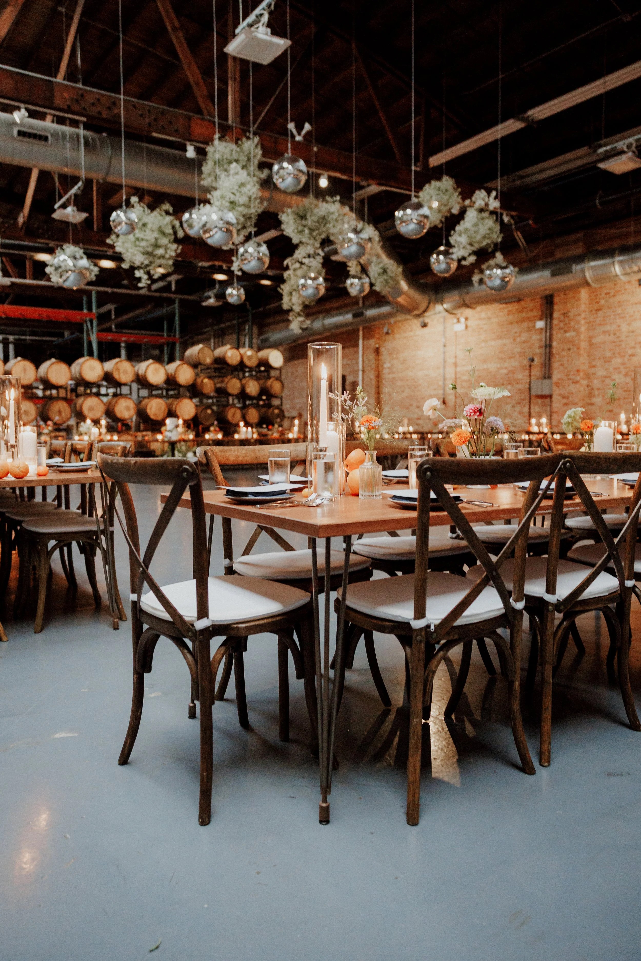 Indoor event space with wooden tables, white chairs, floral centerpieces, candles, and hanging floral decorations with silver ornaments, against a brick wall and wood ceiling with large metal pipes.