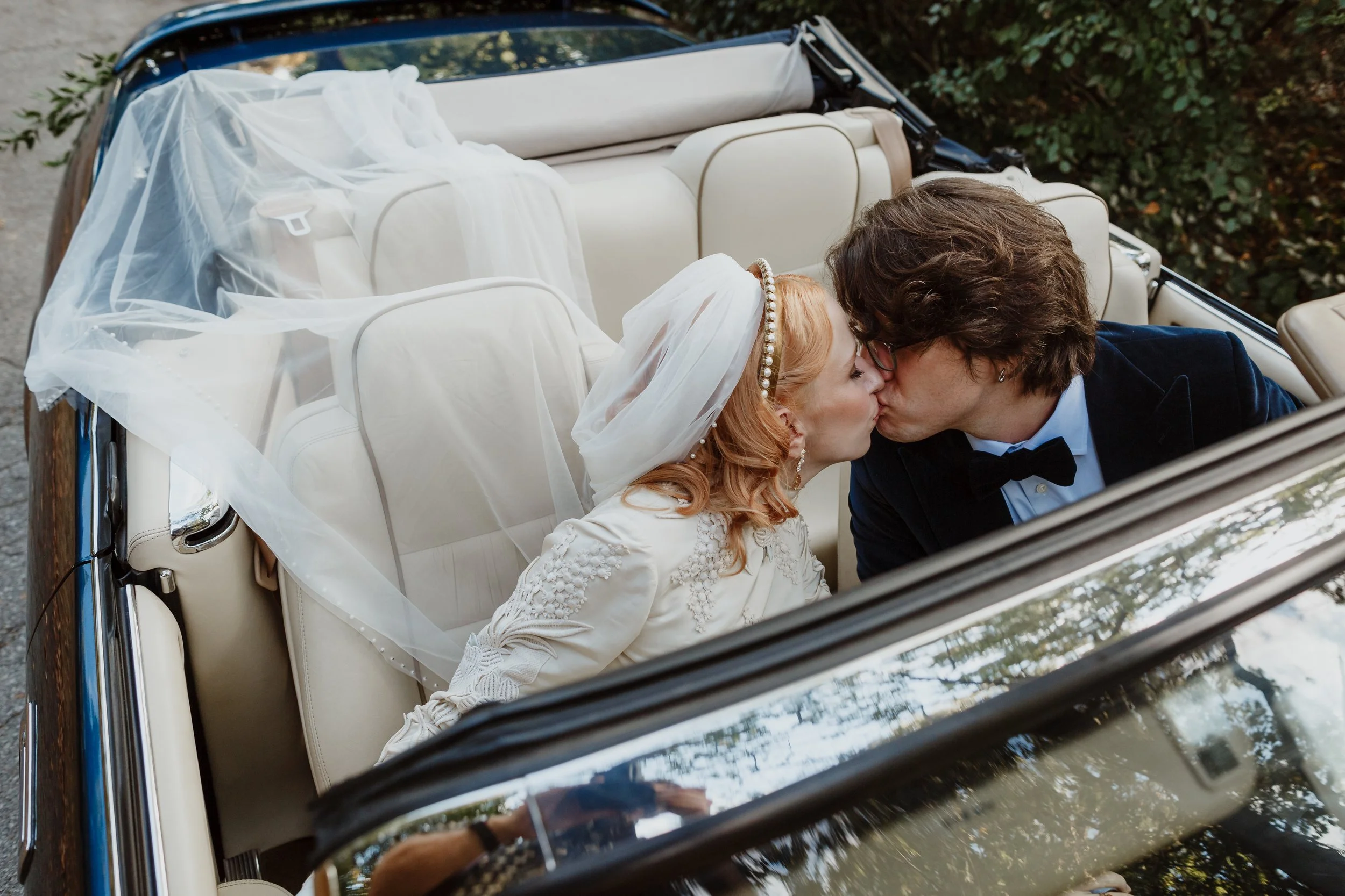 A bride and groom sharing a kiss while sitting in a vintage convertible car decorated for a wedding.