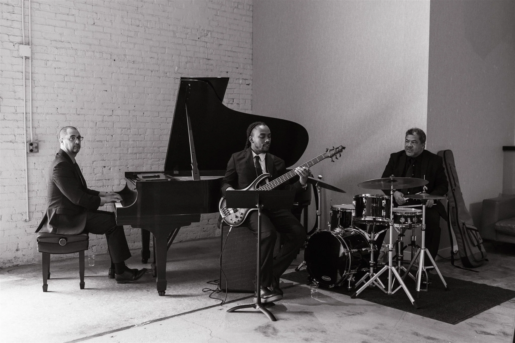 A jazz band performing indoors, featuring a pianist, a bassist, and a drummer, against a brick wall.