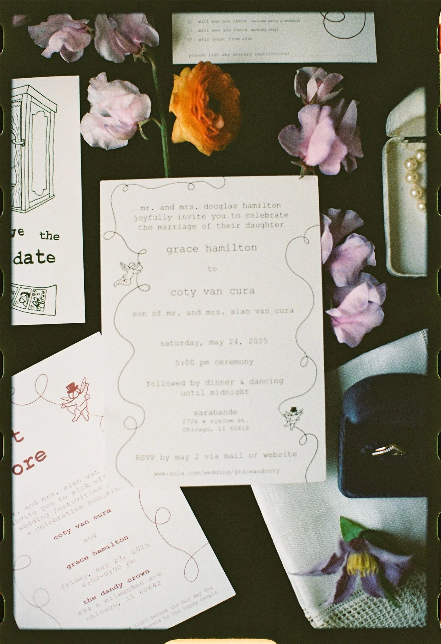 Wedding invitation suite with floral decorations, including purple and orange flowers, and pearl embellishments, placed on a dark surface.