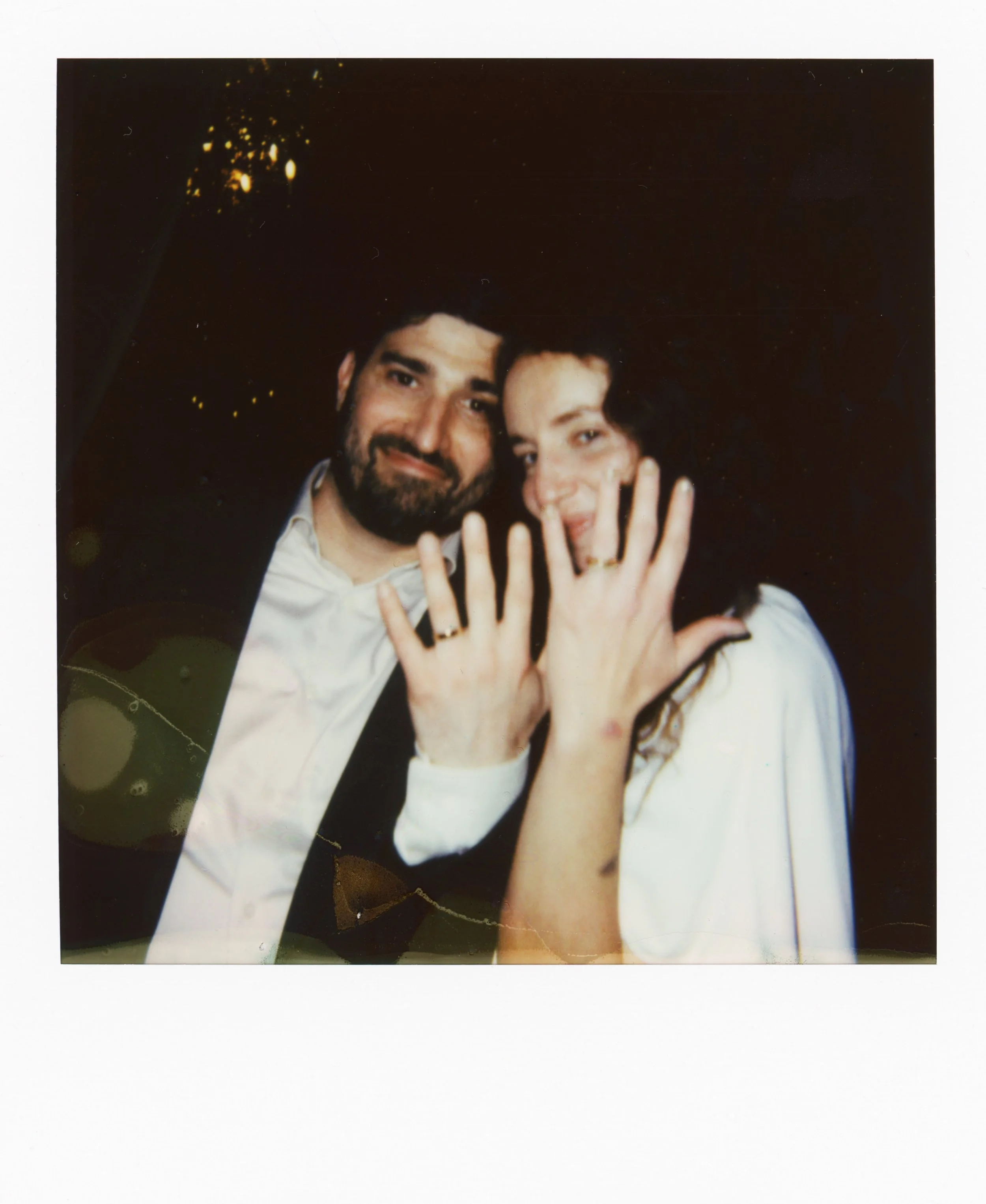A man and woman posing for a photo, both smiling, with the woman showing her hand with a ring on her finger.