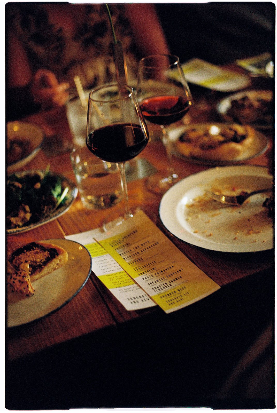 Two glasses of red wine and empty plates on a wooden table with a menu, partially eaten food, and various dishes, suggesting a dinner setting in a restaurant.