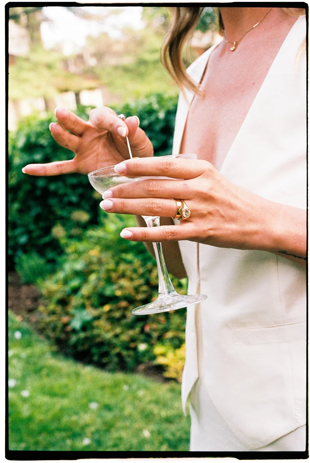 Close-up of a woman's hands holding a cocktail glass outdoors, seemingly preparing to add a garnish or stir, with a blurred garden background.