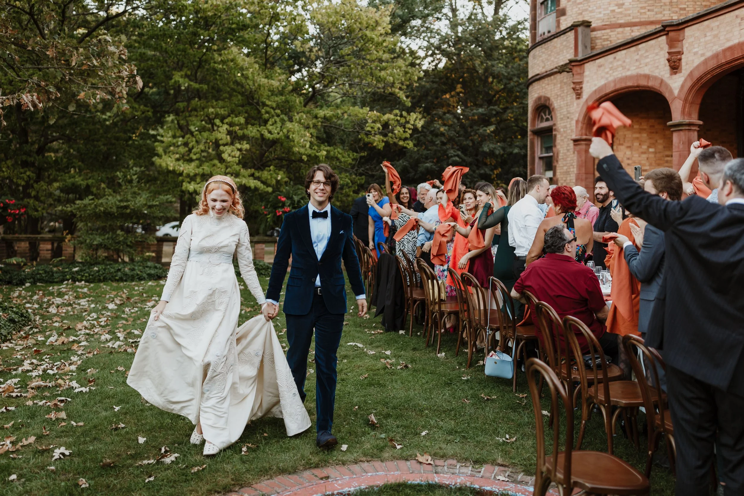 A bride and groom holding hands walking through a wedding reception outdoors, smiling. Guests are seated at long tables, some waving orange napkins, with a brick building and green trees in the background.