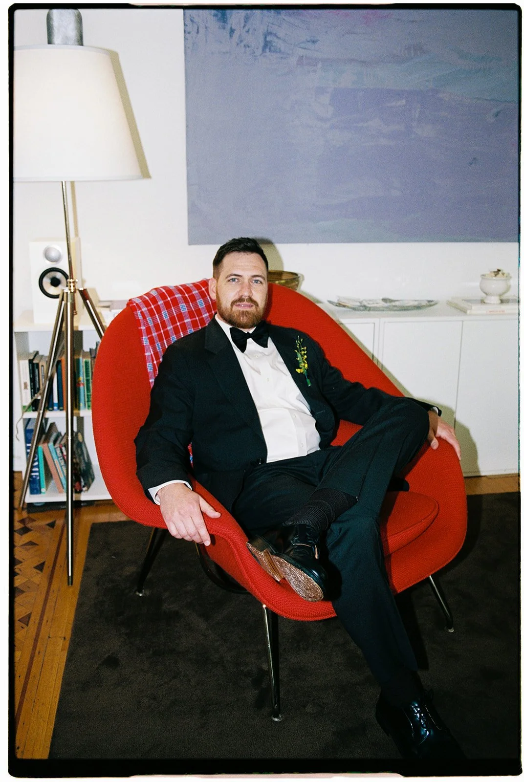 A man in a tuxedo with a floral boutonniere sitting in a red modern lounge chair in a living room, with a black coat, white shirt, and black shoes, surrounded by books and modern decor.